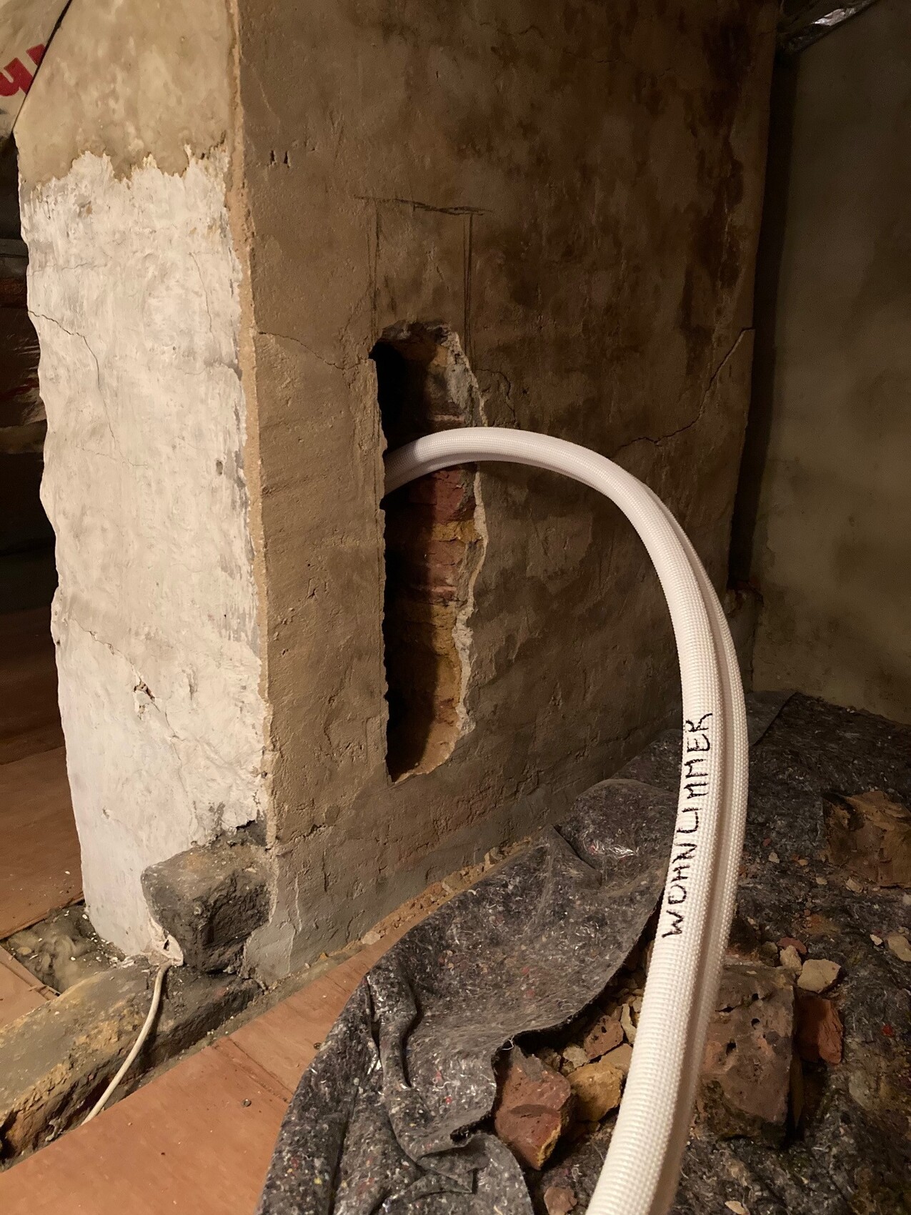 Basement wall with a hole in the concrete surface; white flexible hose runs through the opening, construction debris nearby.