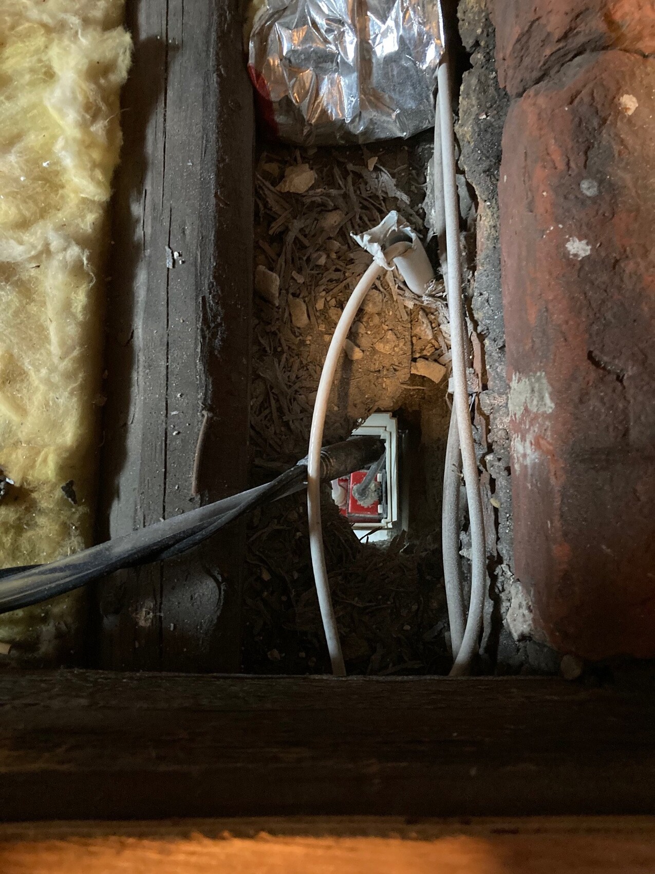 Exposed cables in a dusty wall opening between brick and wood; insulation on the left, red interior section visible.