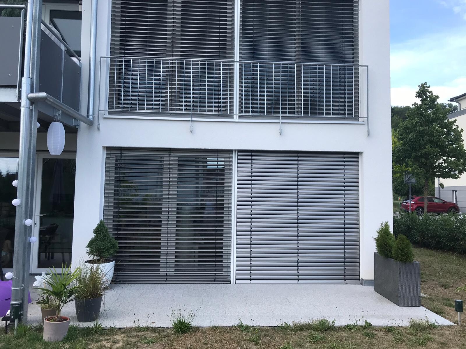 Modern white house facade with large windows, blinds, balcony, and string lights on the left