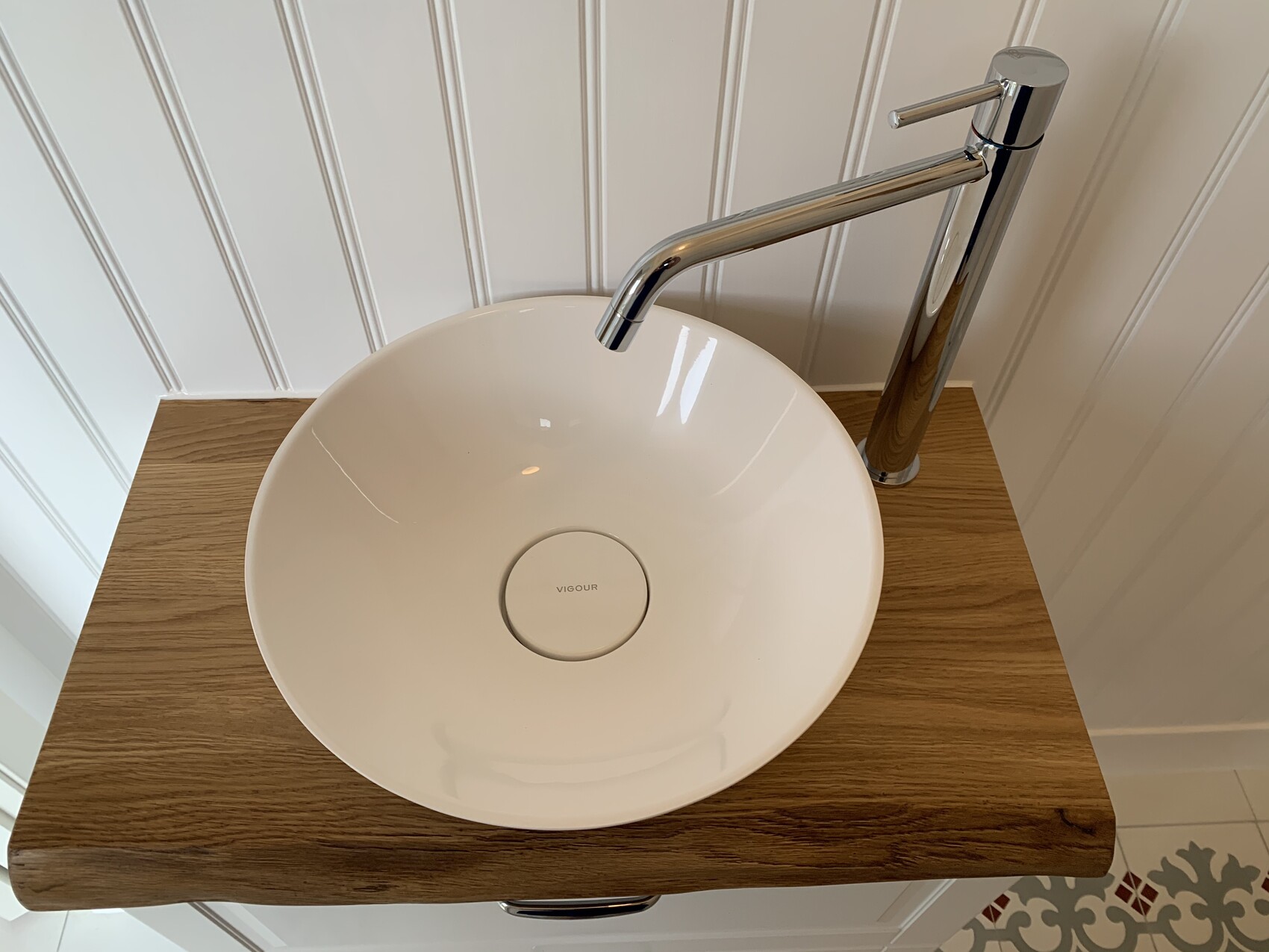 Round, white sink on wooden surface with chrome faucet in front of white paneled wall.