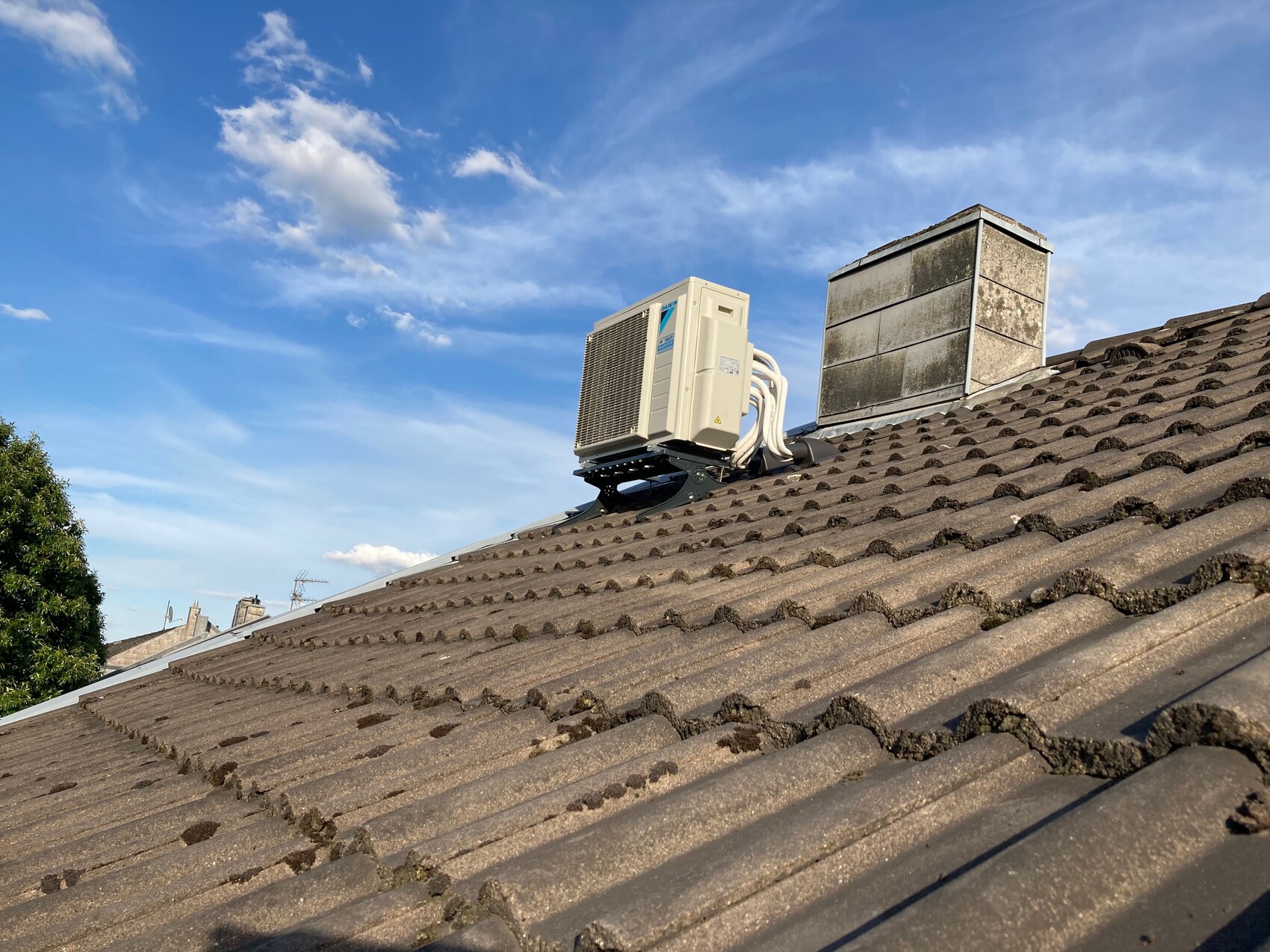 Outdoor air conditioning unit on a sloped tiled roof next to a chimney; blue sky.