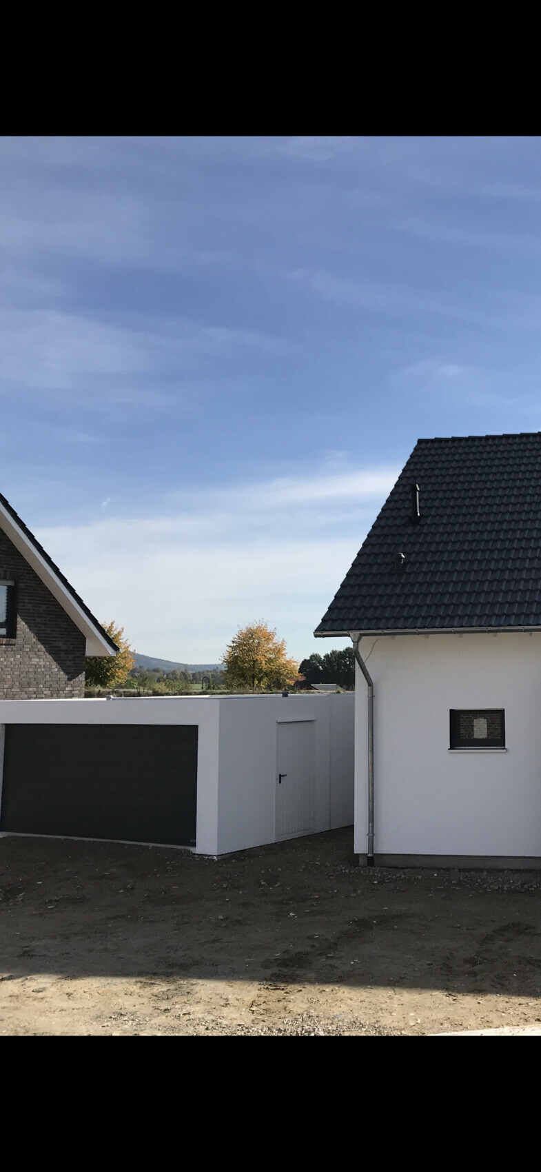 Moderne weiße Häuser mit dunklem Dach und Garage vor blauem Himmel, Herbstbäume im Hintergrund