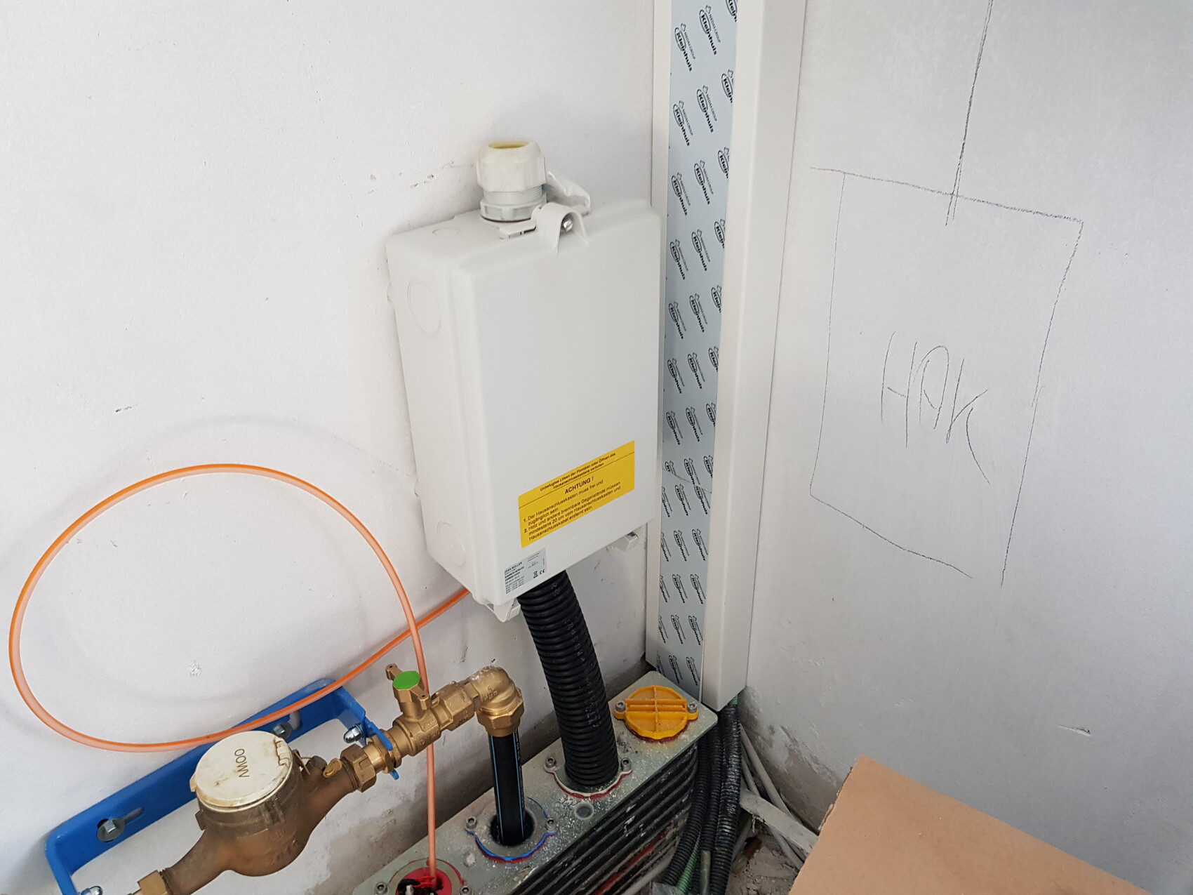 White installation box on the wall, orange hose, brass valve, and cables in the technical room.