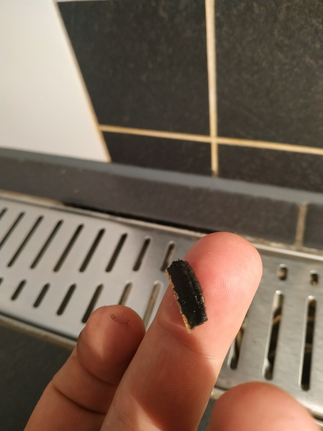 Hand hält kleines schwarzes Kachelstück vor dunkler Badfliese neben Duschrinnenabdeckung