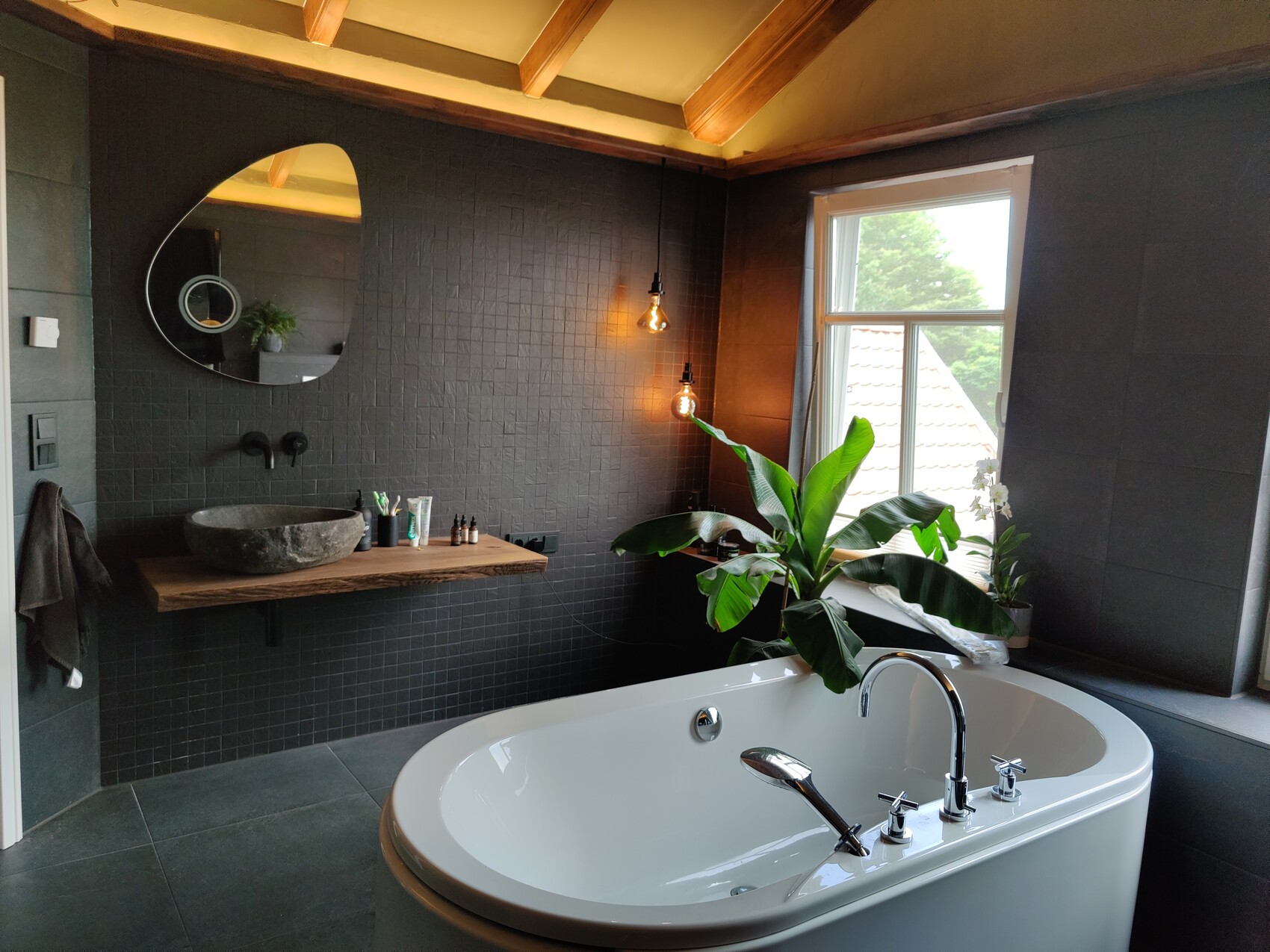 Freestanding bathtub in front of dark tiles; wooden shelf with stone basin, round mirror, plant.