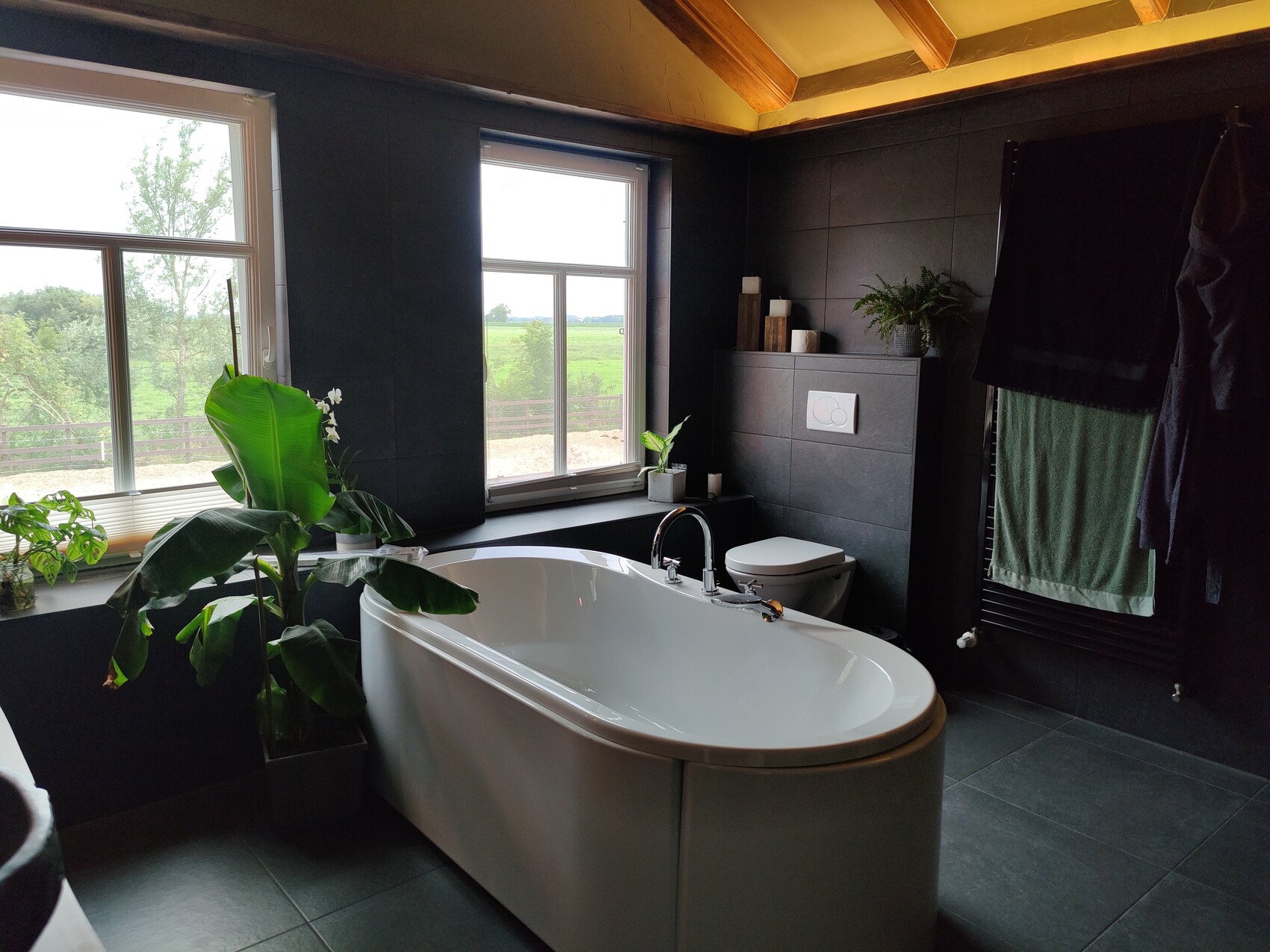 Freestanding white bathtub in front of dark tiles, large windows, and green plants.