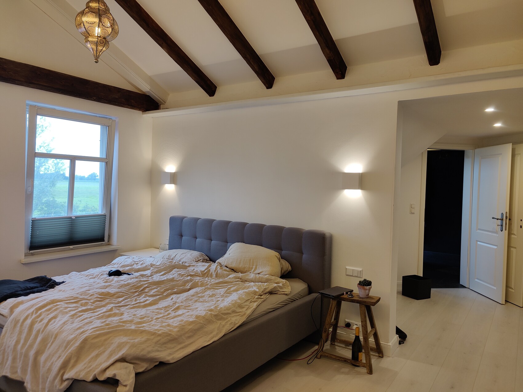Bedroom with gray upholstered bed, wooden beam ceiling, window and door to hallway.