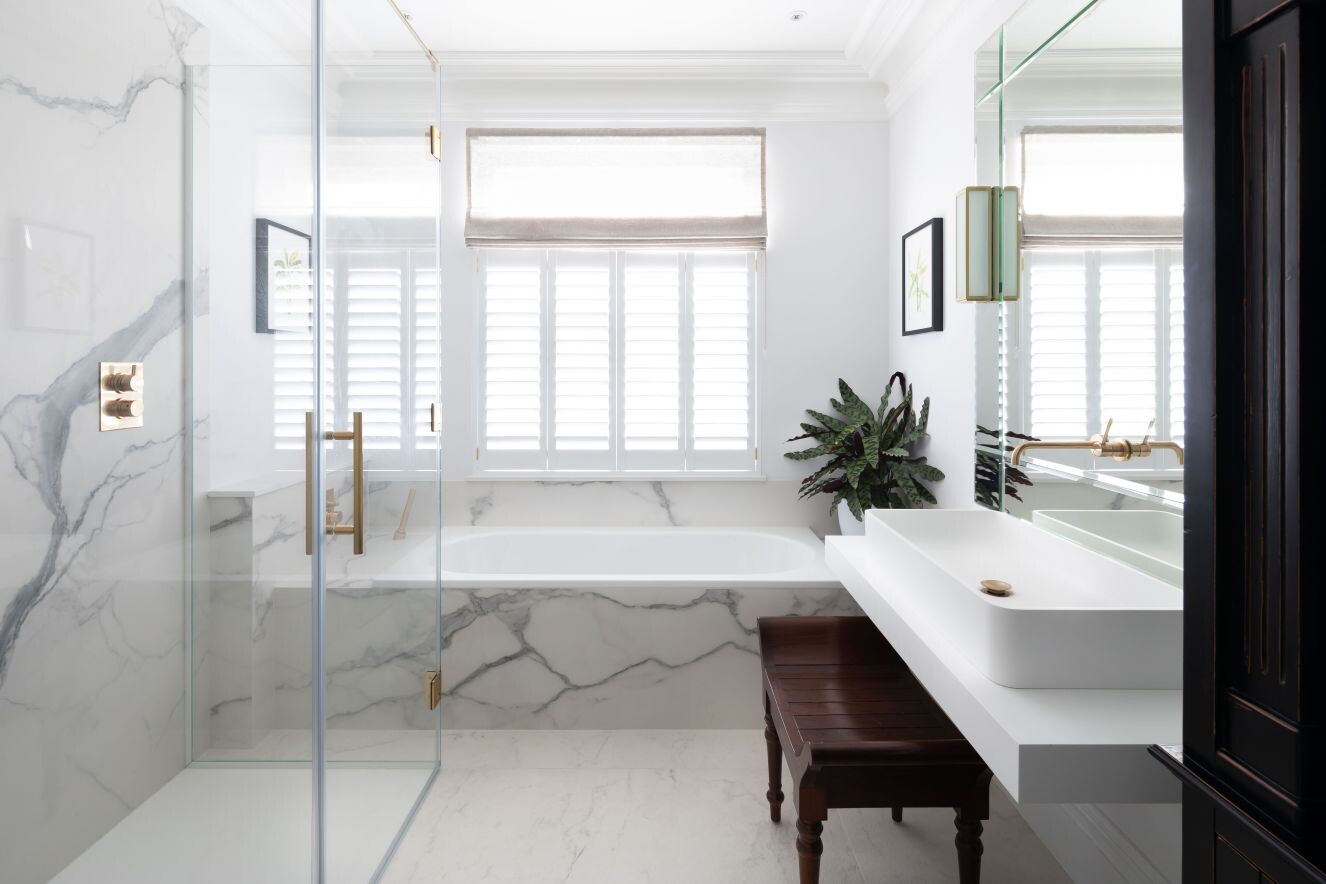 Bright, modern bathroom with marble walls, glass shower, bathtub, shutters, and plant.