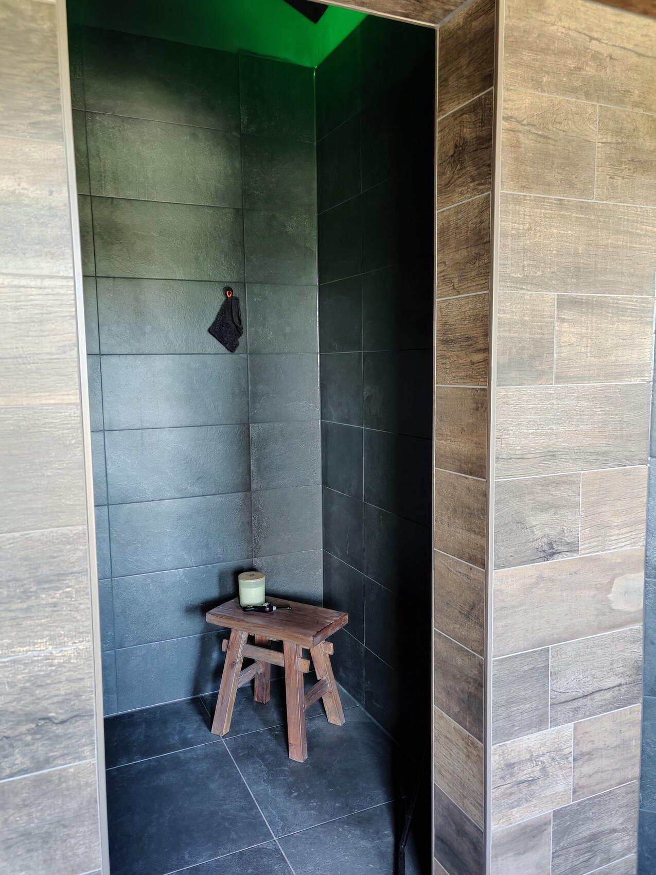 Dark shower area with gray tiles, green light above, small wooden chair with candle on it