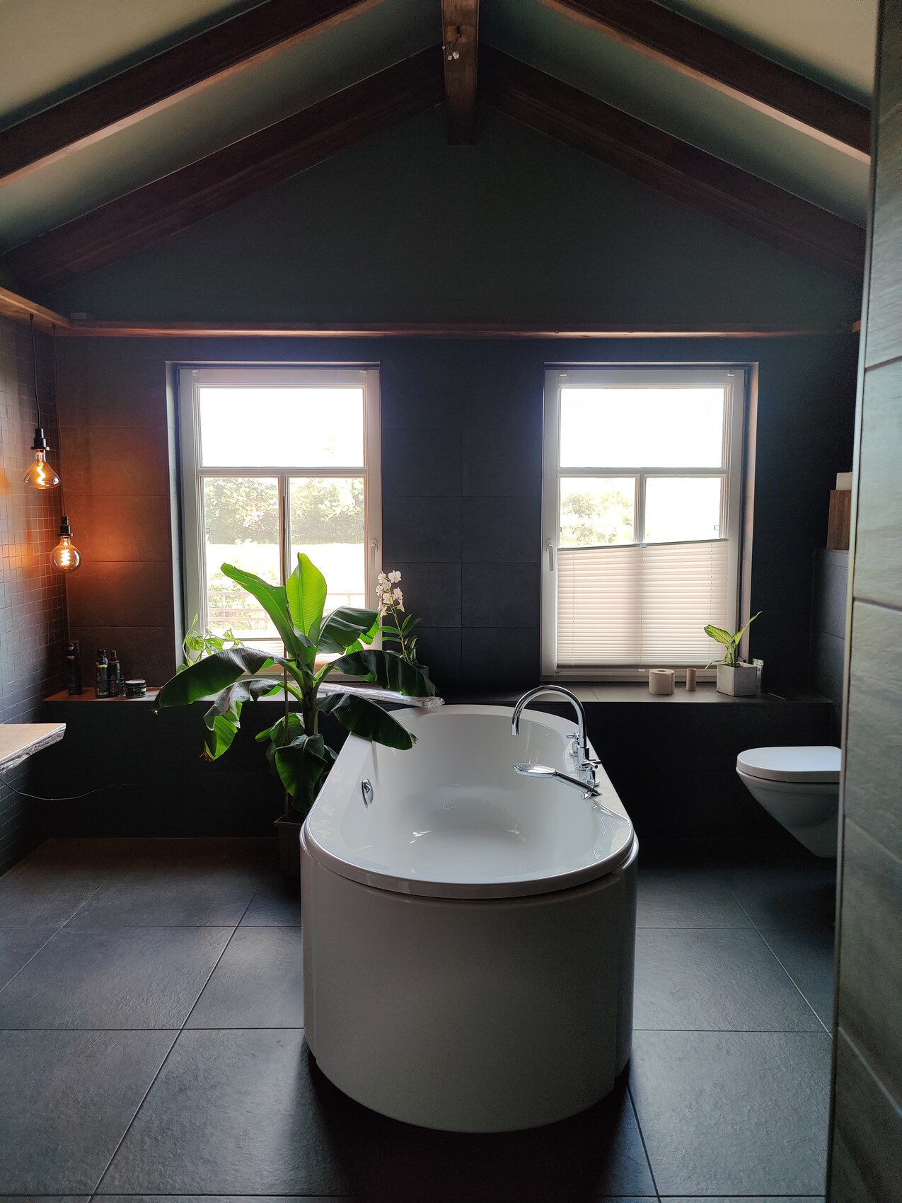 Bright bathroom with freestanding bathtub, dark tiles, two windows, and plants.