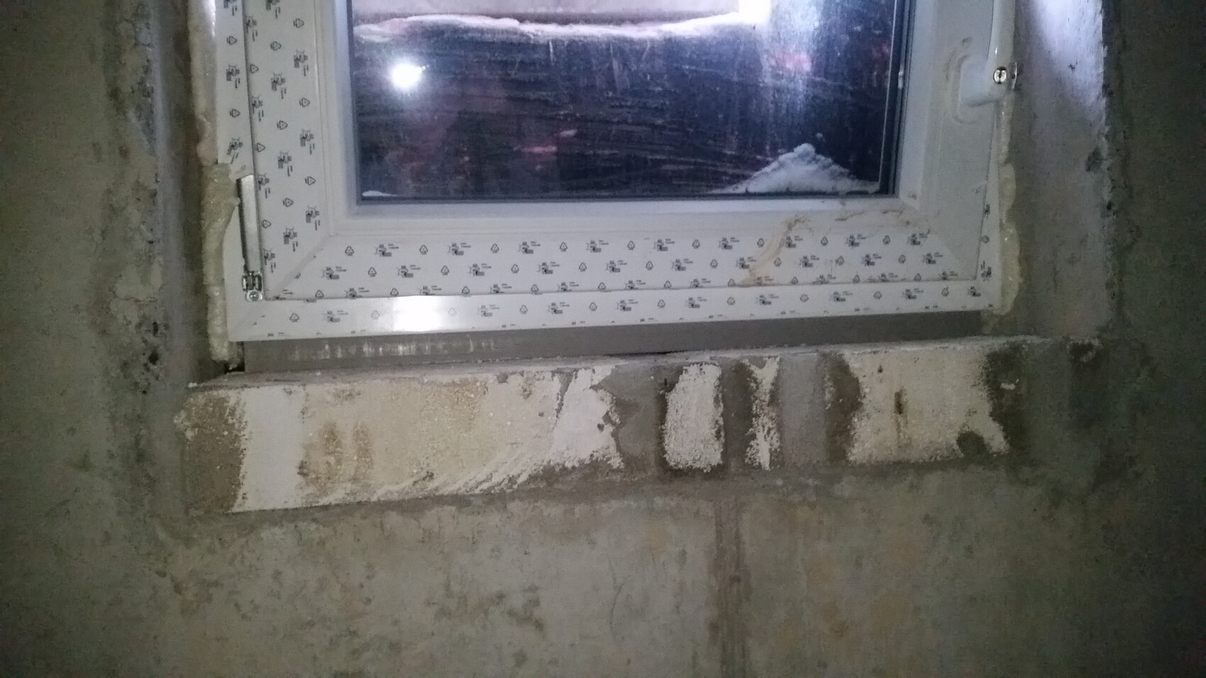 Kellerfenster in roher Betonwand; weißer Rahmen, Putzreste und Feuchtigkeitsspuren.