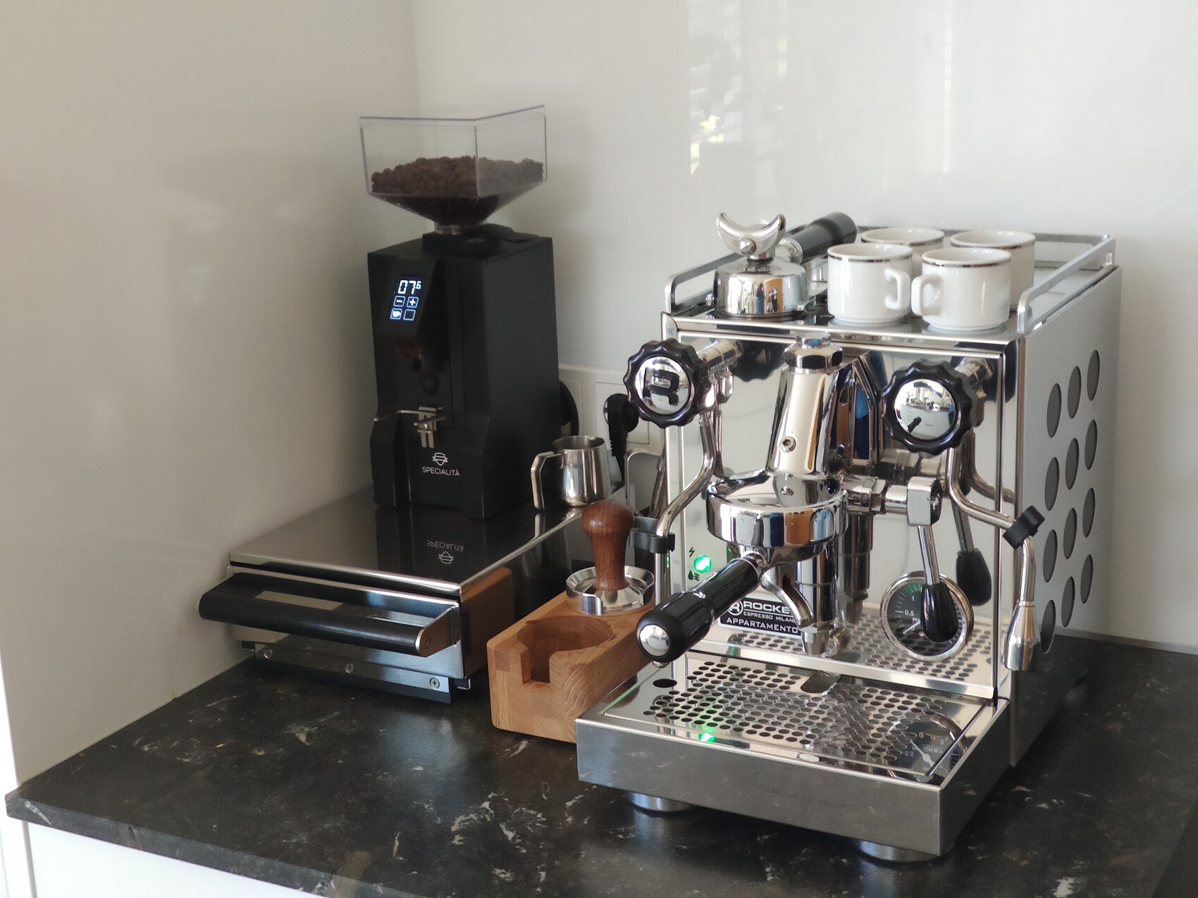 Espresso machine made of stainless steel with two group heads, cups on top, grinder on the left, and tamper on the counter.