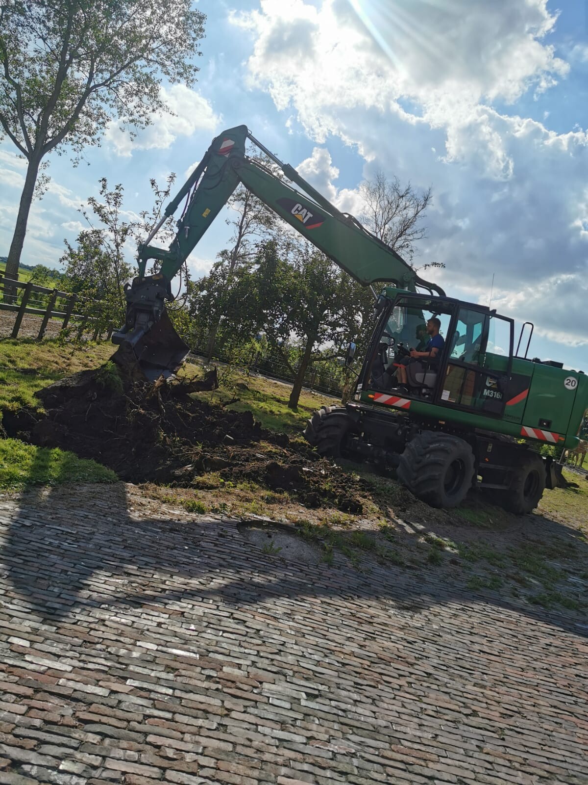 Green CAT excavator with long arm digging soil next to a cobblestone path; operator in the cabin.