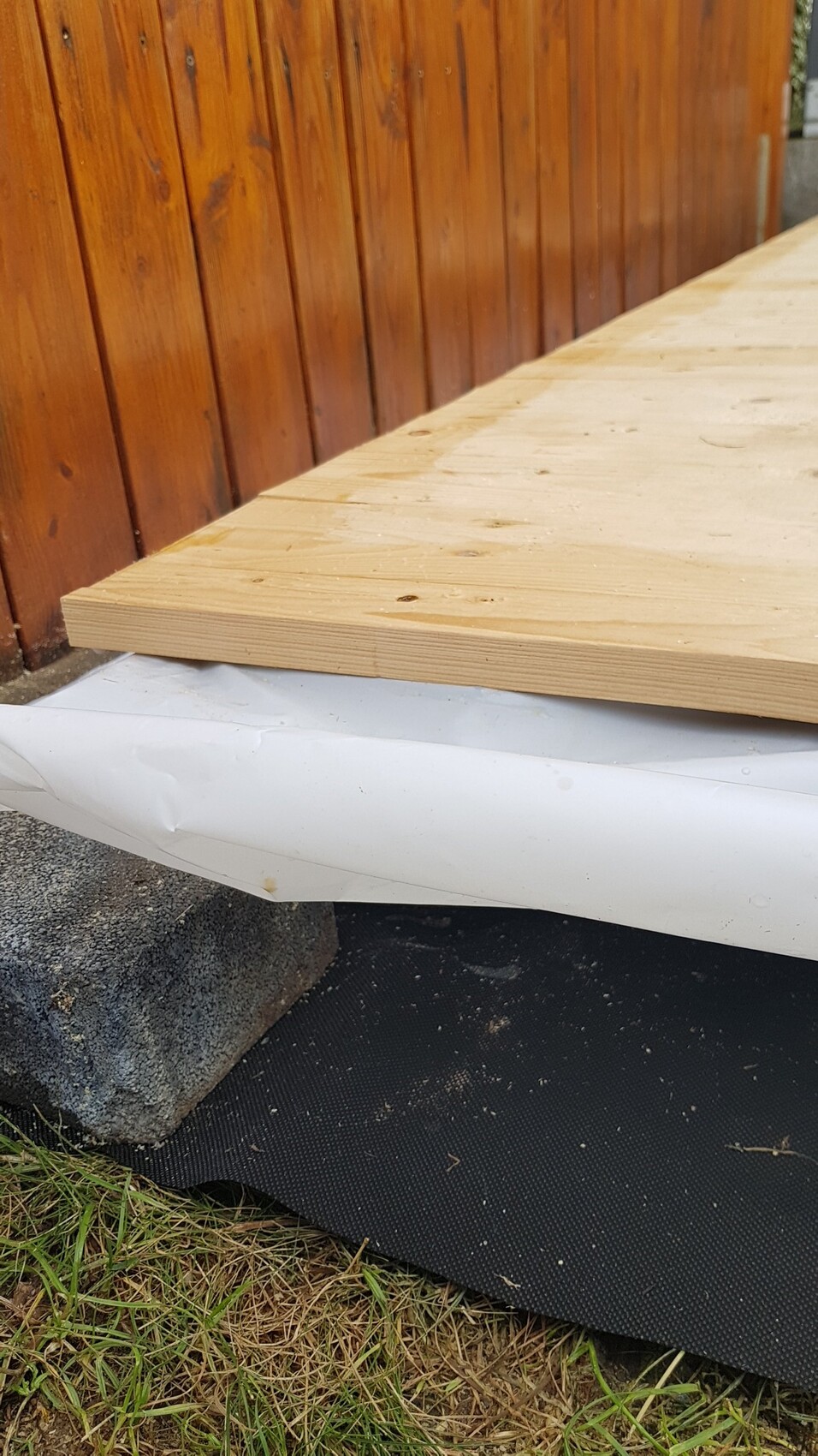 A wooden board lies on a white construction membrane over the foundation next to a vertical wooden wall.