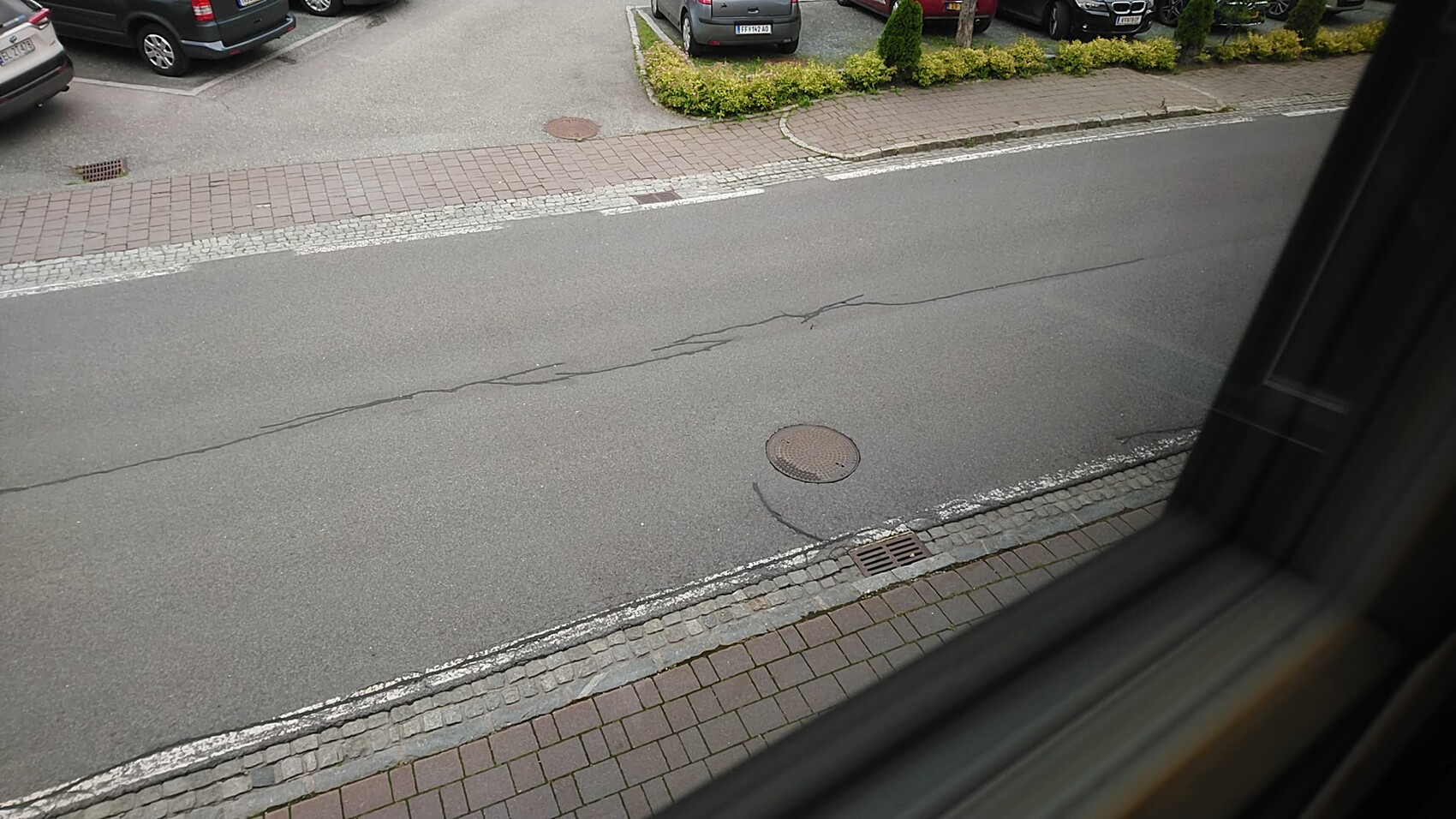 View from window onto a street with parked cars, sidewalk, manhole cover and cracks in the asphalt.