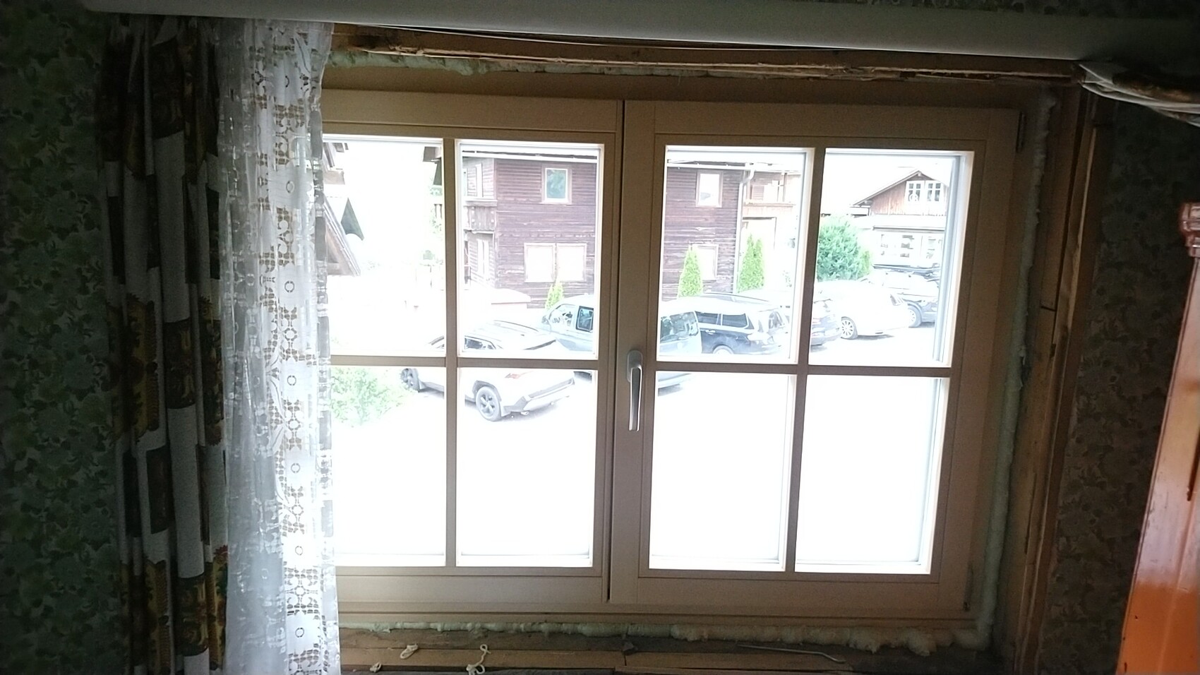 Window with light frame, view outside to parked cars in front of wooden houses; left peak curtains.