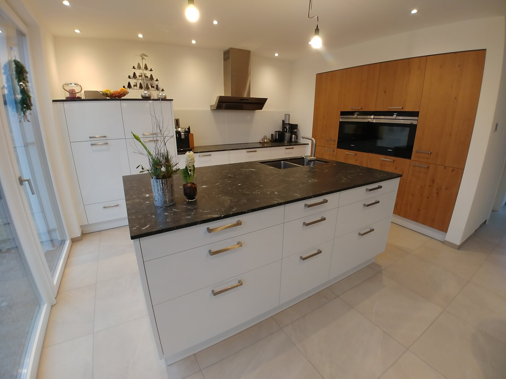 Modern kitchen with large island, black stone countertop, sink, oven in wooden cabinet wall, stainless steel.