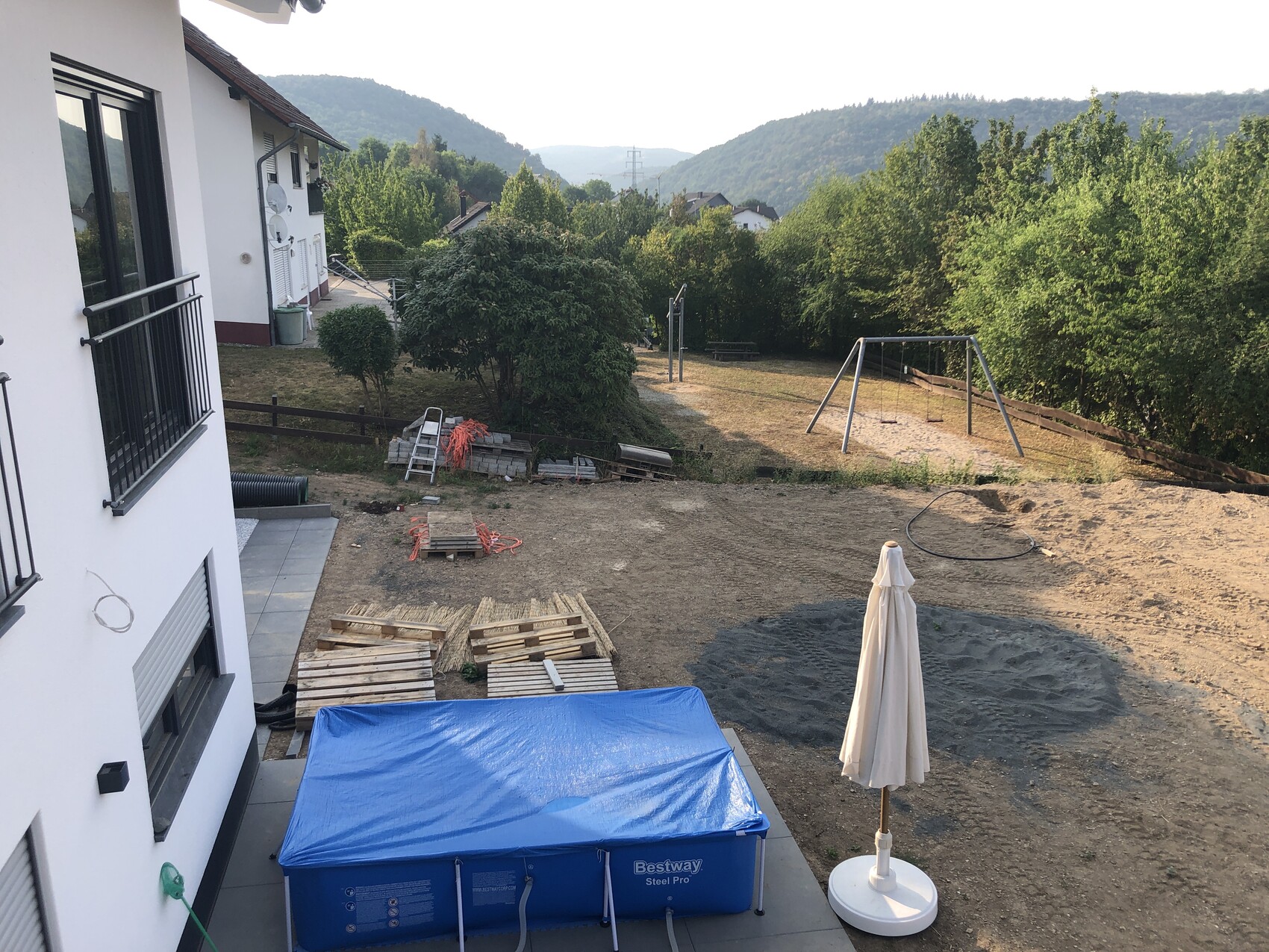 Garten- und Baufläche neben Wohnhaus: Paletten, blaue Abdeckplane, Schaukel, Bäume im Hintergrund.
