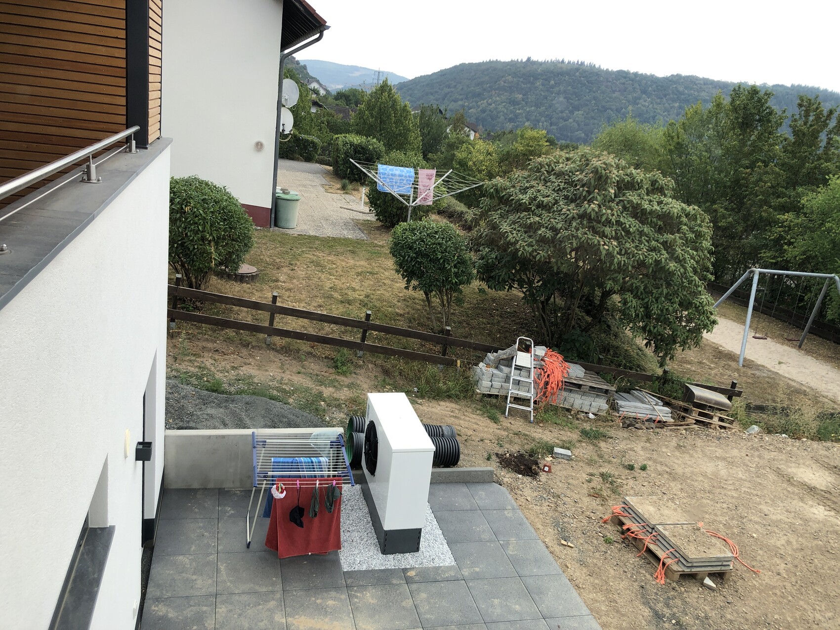 Terrace of a house with a drying rack, garden, trees, playground, and mountain landscape in the background