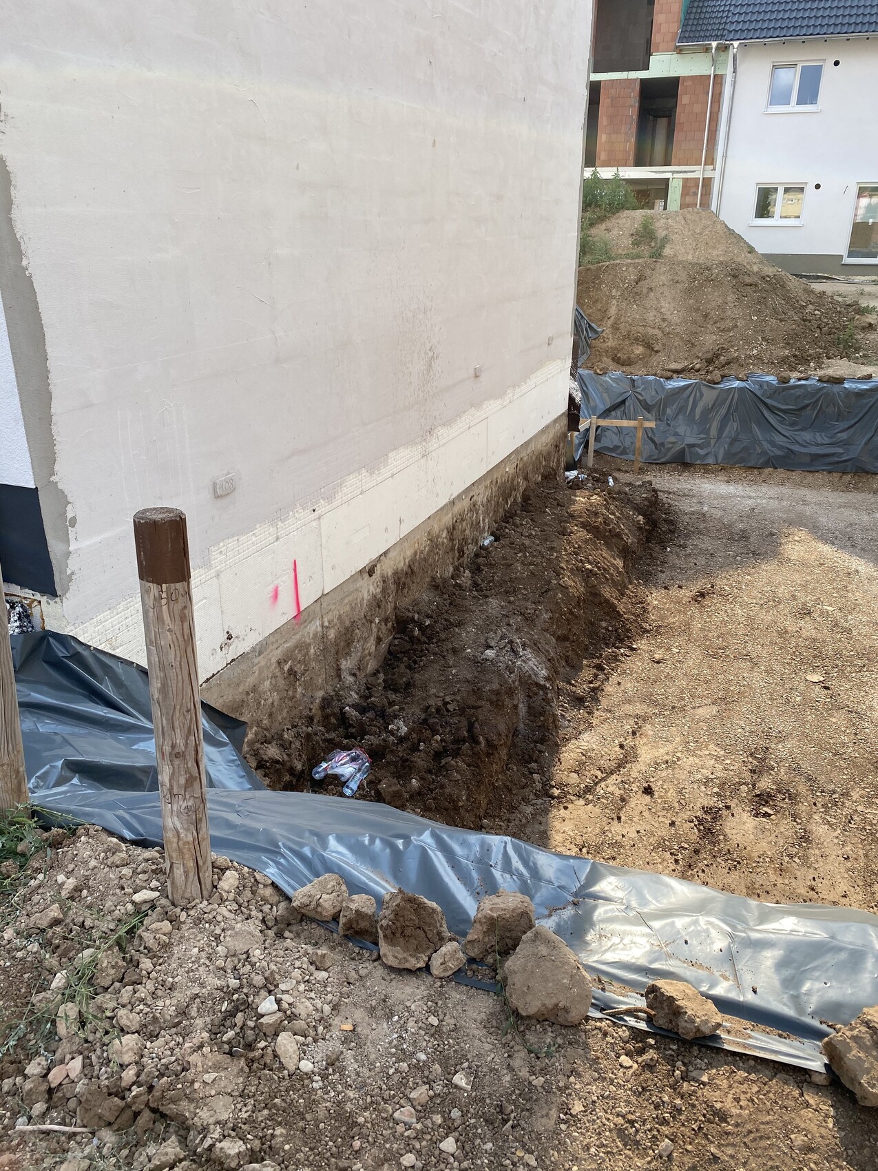 Construction site: trench next to white building wall, earth excavation, black foil, wooden posts.