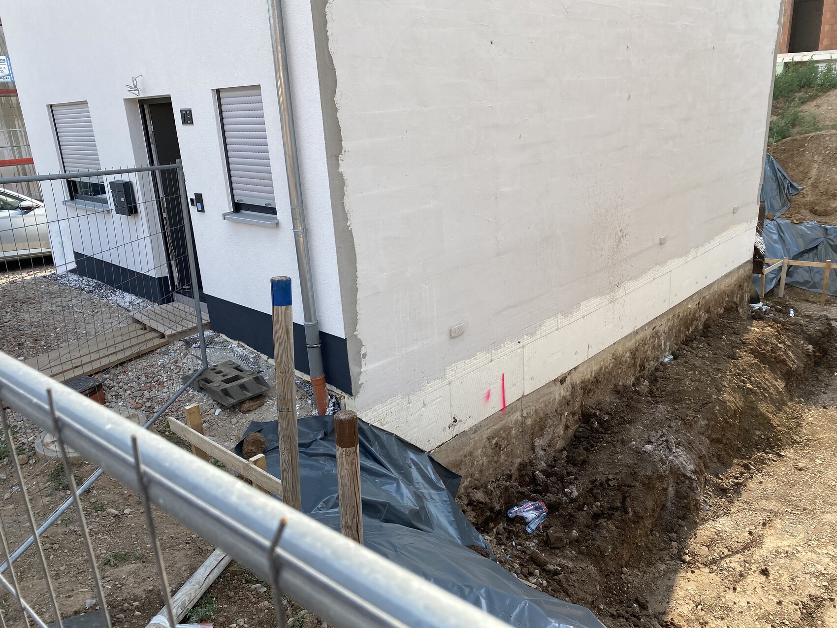 Construction site next to white house: trench at foundation, construction fence in the foreground.