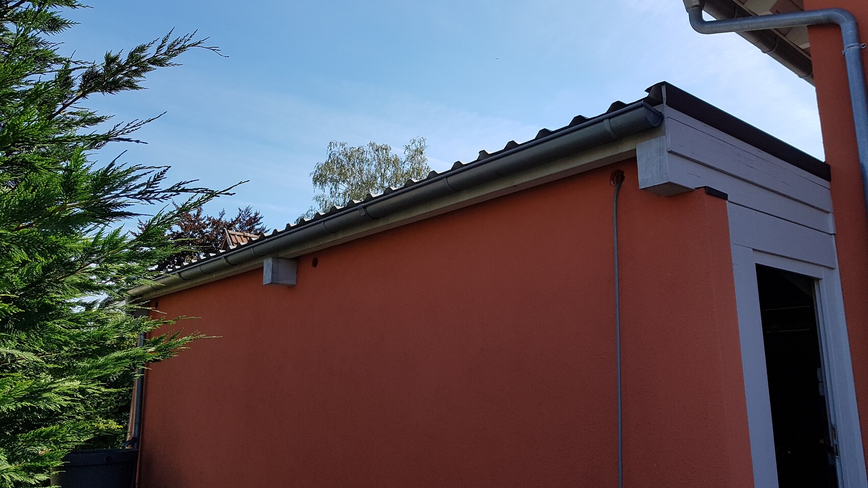 Orange building wall with white wooden cladding, open door, blue sky, trees on the left.