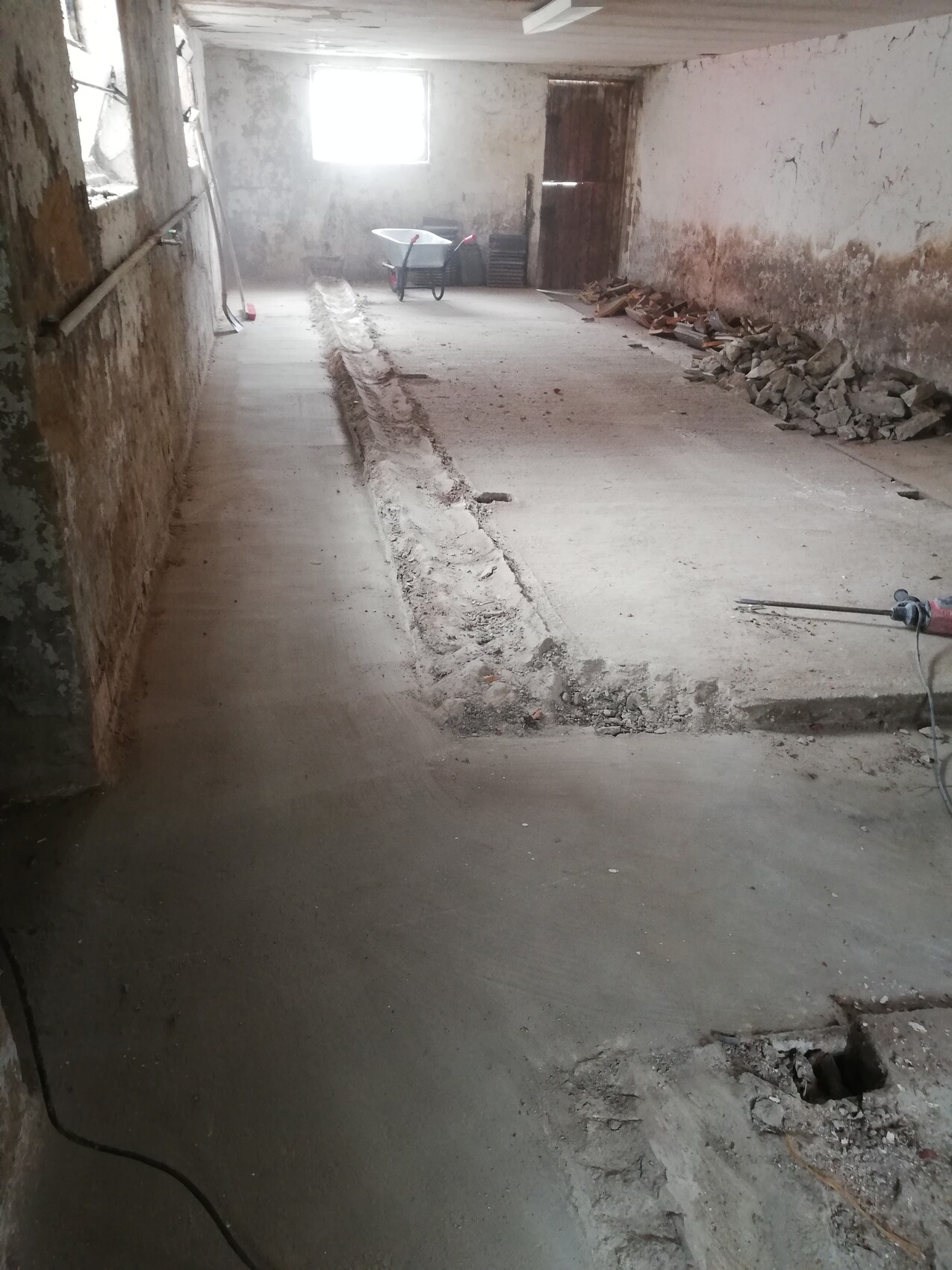 Basement construction site: rough concrete floor, trench in the middle, debris on the right, wheelbarrow in the background.