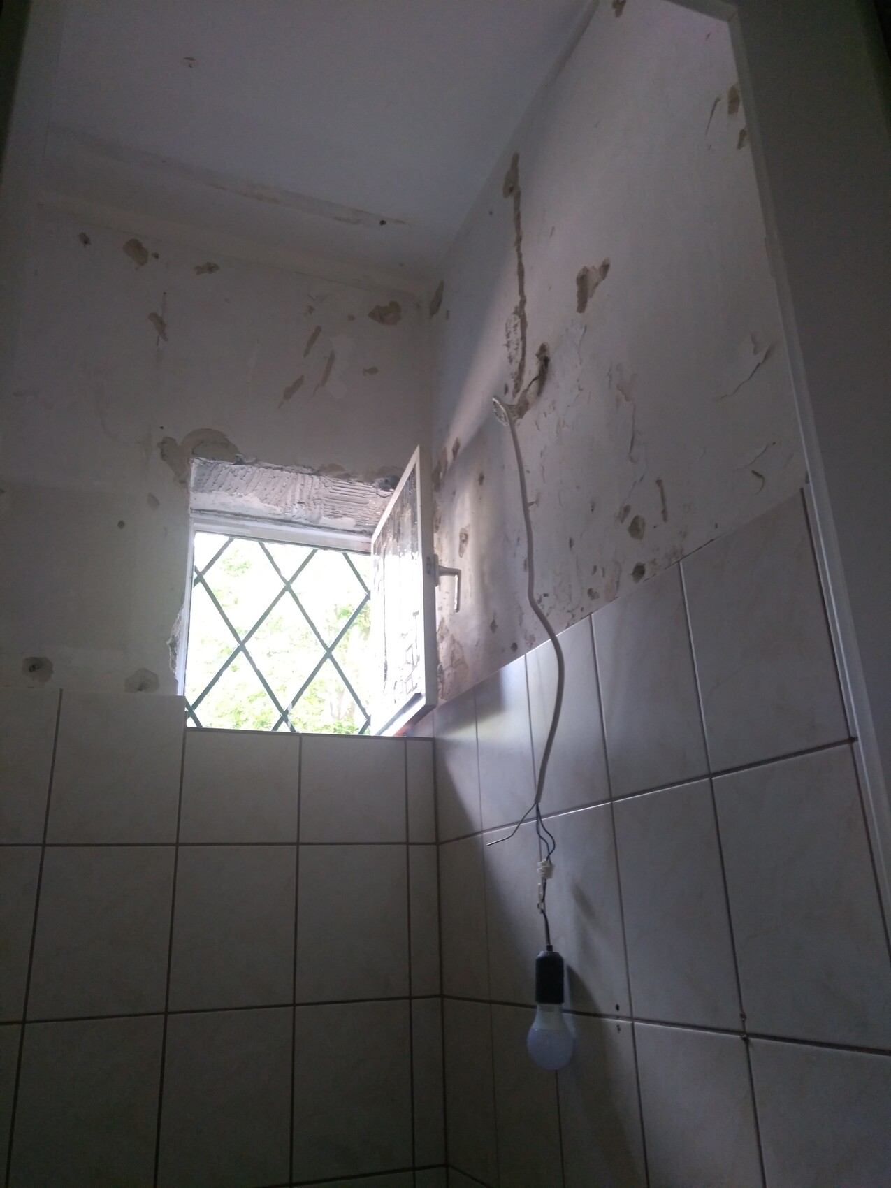 Small bathroom with peeling walls, tiles, open window with grate and light bulb.