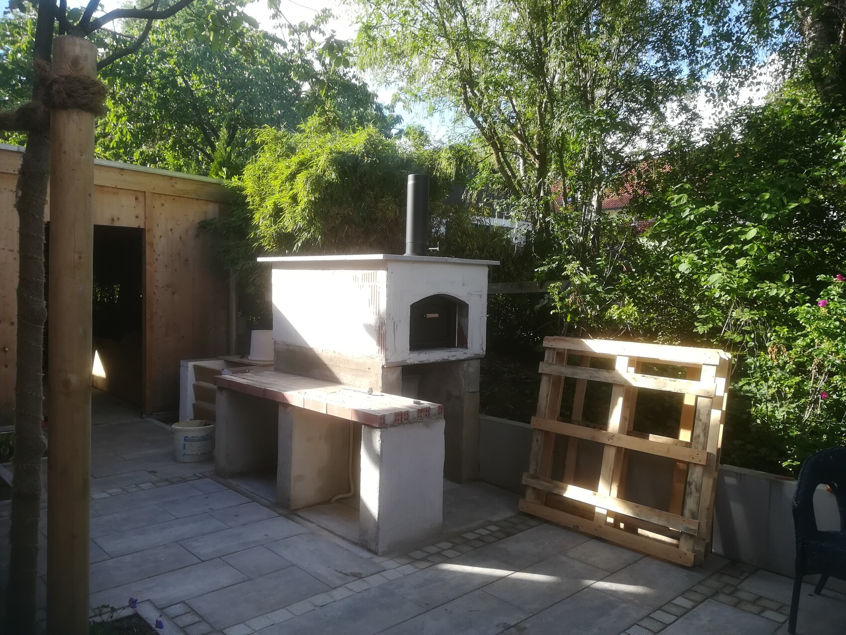 Garden area with white exterior oven on stone base, wooden bench and pallets in front.