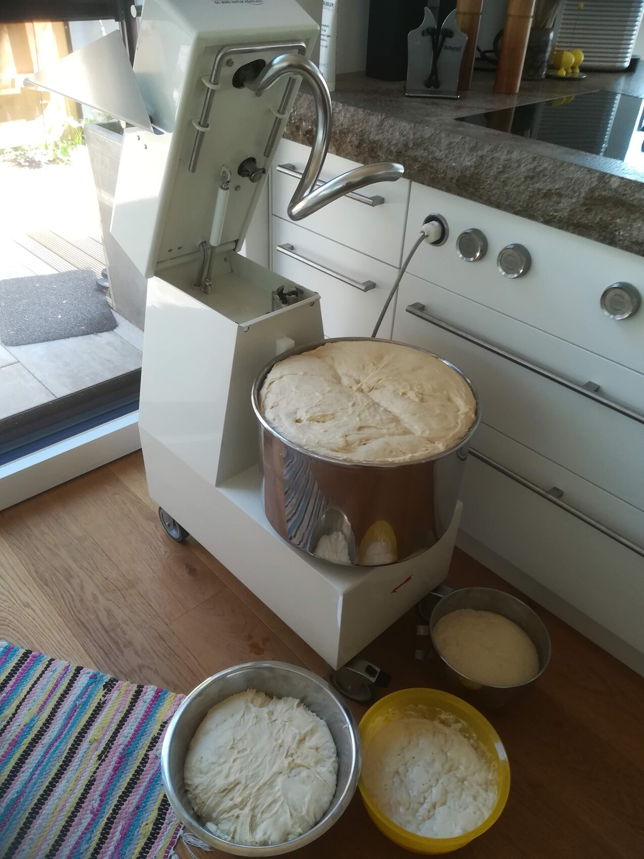 Large white kitchen mixer with open lid; several dough bowls with yeast dough next to it.