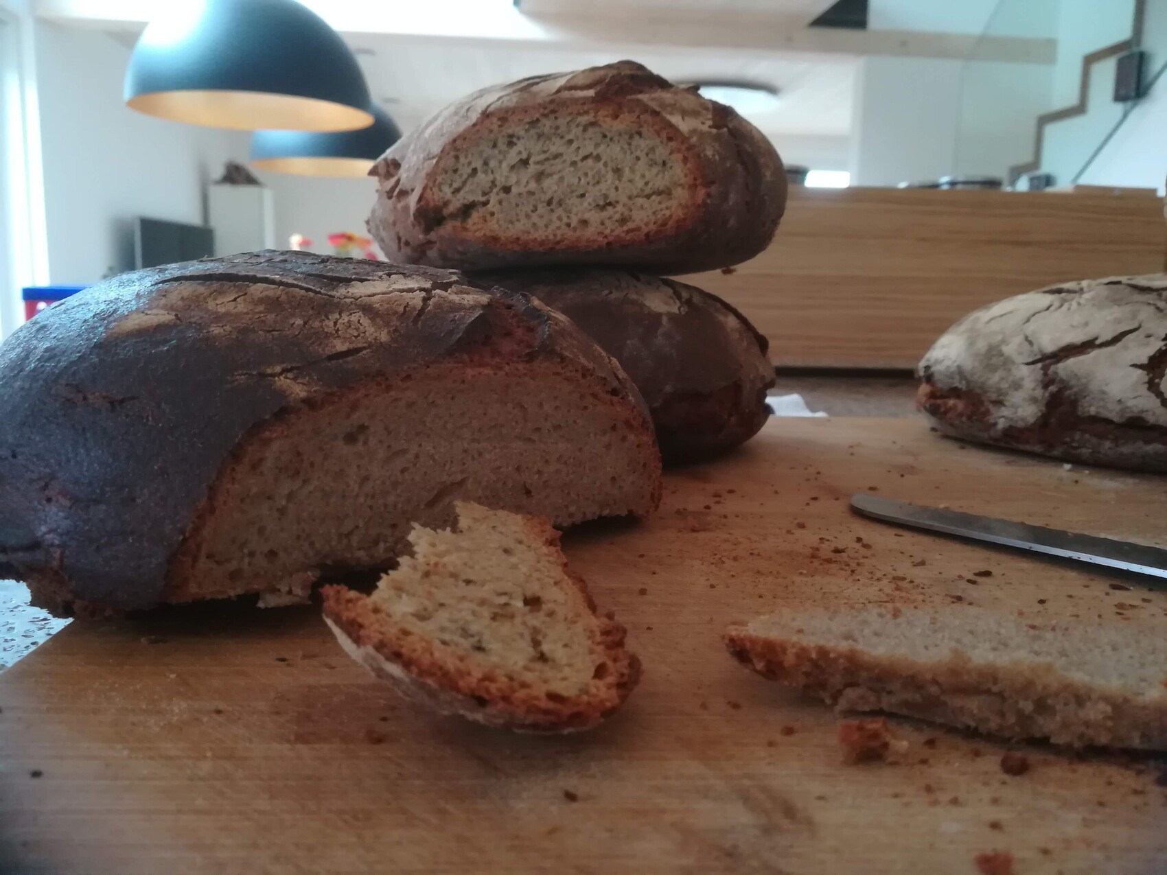 Dark bread on a wooden board; a slice broken off with a knife beside it; kitchen background.