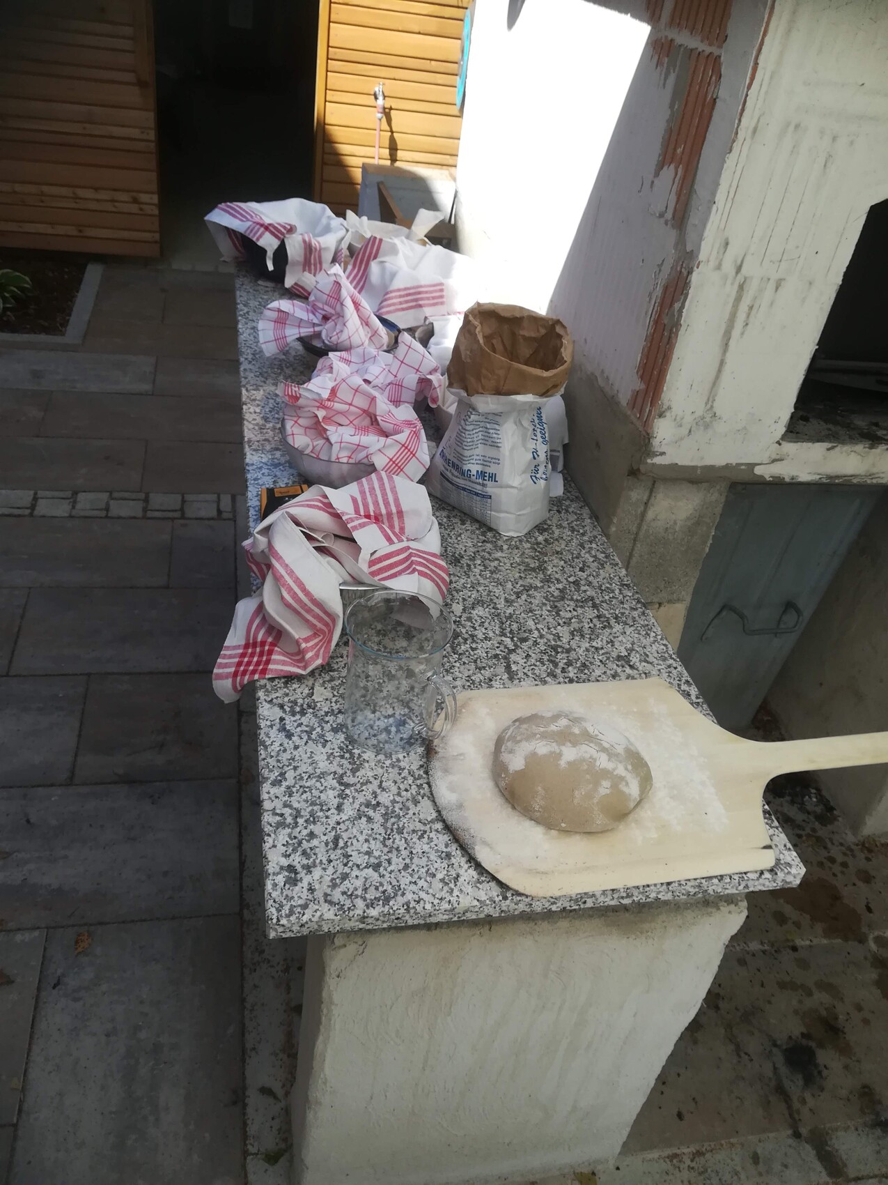 A dough ball on a wooden peel next to bags of flour on a granite slab outdoors.