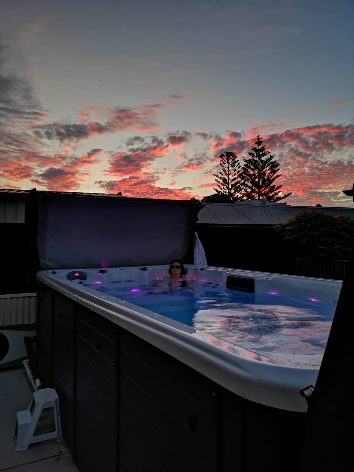 Person relaxing in an illuminated outdoor hot tub with purple light at sunset.