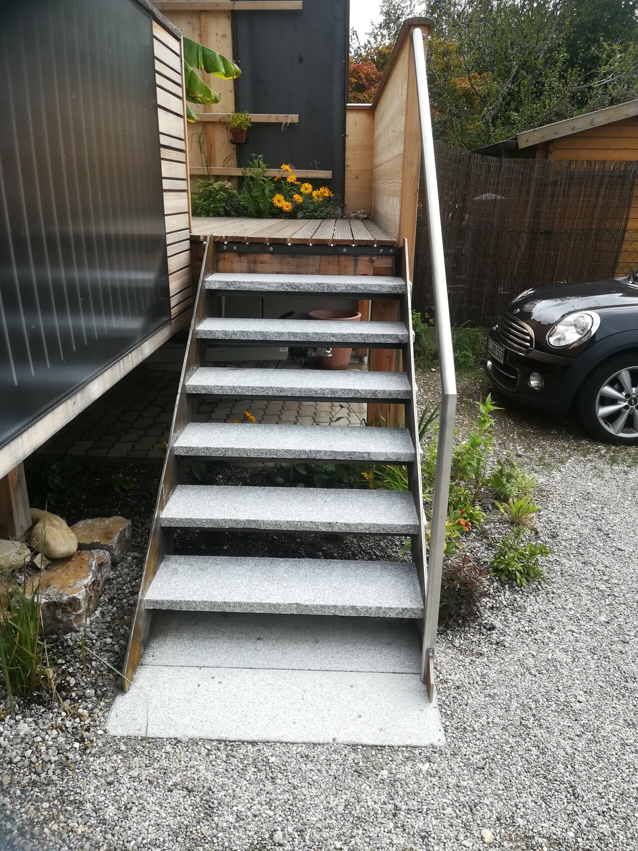 Outdoor metal steps leading to a wooden platform; yellow flowers, parked car nearby.
