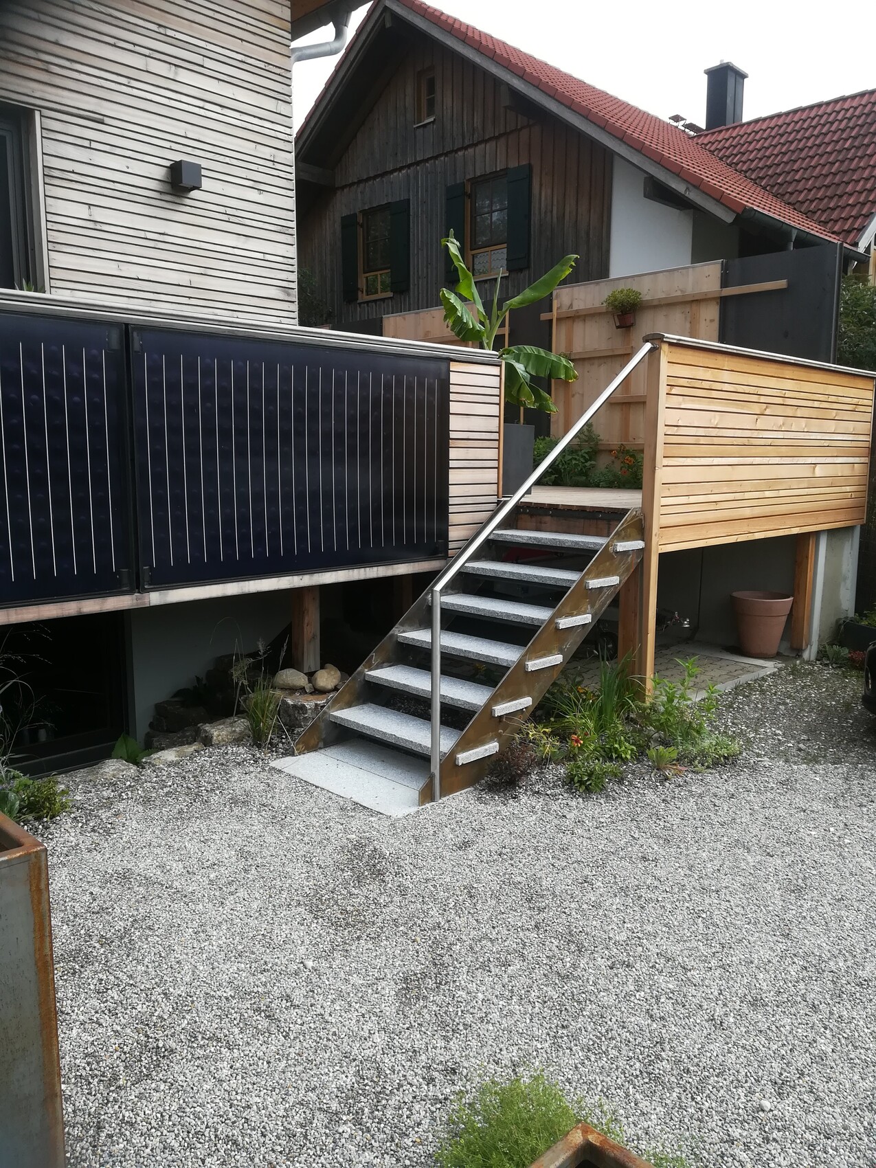 Outdoor metal staircase leading to a wooden terrace with railing beside the house; gravel ground and banana.
