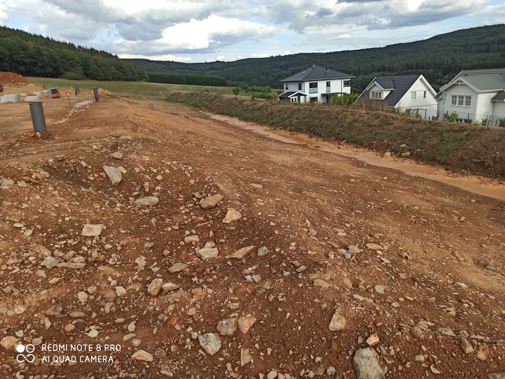 Construction site with soil, gravel, and foundation shells; residential houses in the background.