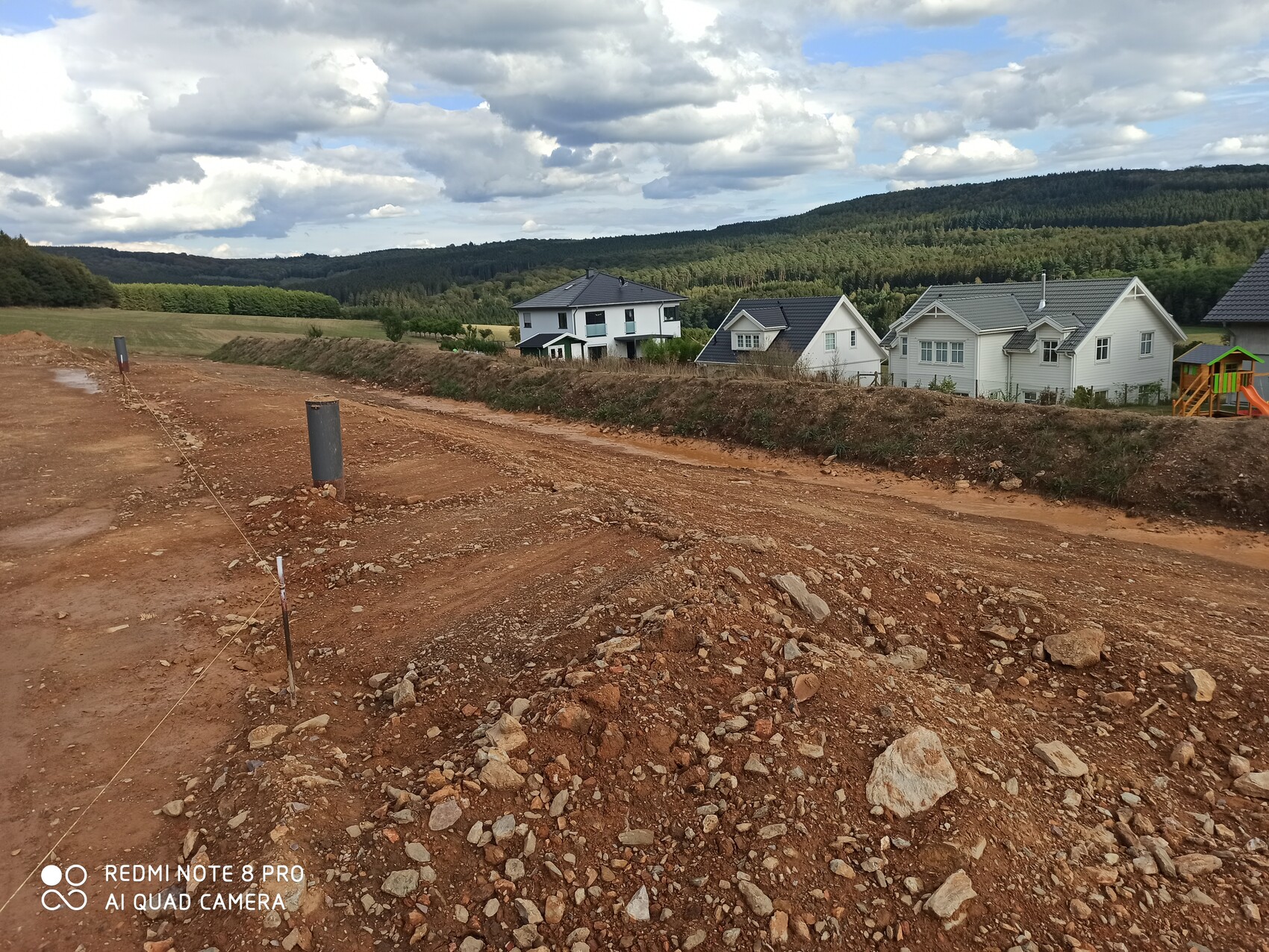 Construction site with red soil and gravel, white houses on a slope, forest in the background.