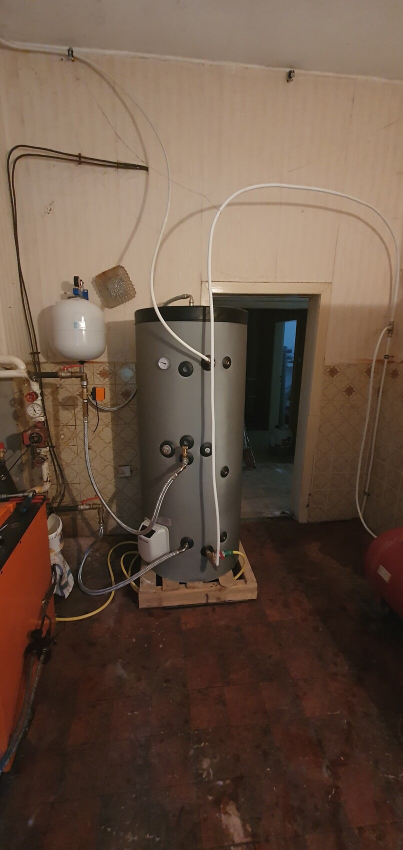 Large cylindrical hot water storage tank with pipes and pressure gauges in the basement.