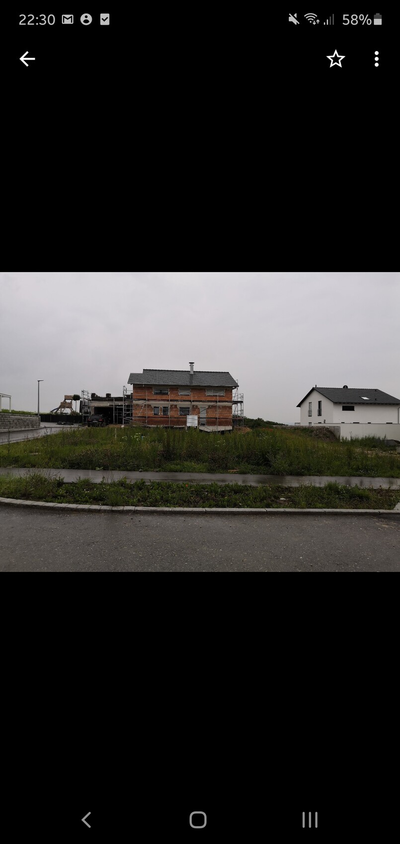 Zweistöckiges Haus im Bau mit Gerüst; daneben ein fertiges weißes Haus, Baustelle auf Grasfläche.