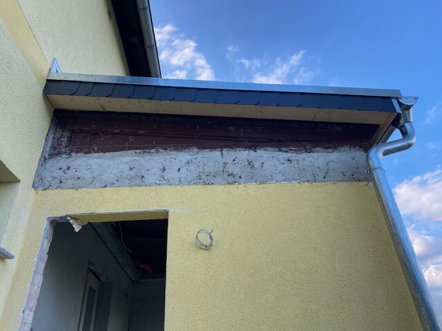 Gelbe Hausfassade mit offenem Eingang, freigelegtem Putz oben, Fallrohr rechts, blauer Himmel