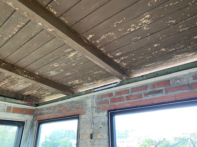 Unverputzter Innenraum: Holzbalkendecke, bröckelnde Decke, Backsteinwände, zwei Fenster, Kabel an Wand.