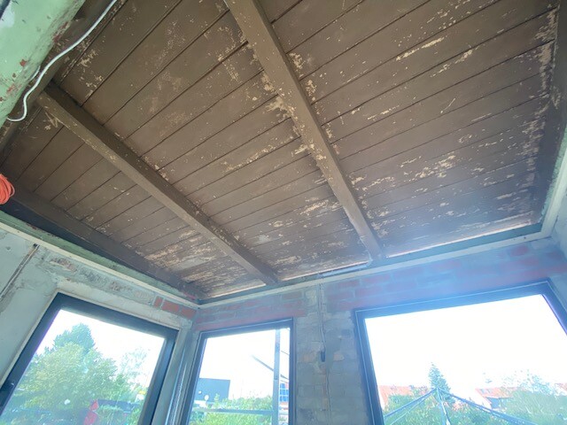 Unbearbeiteter Innenraum: Holzbalkendecke, rohe Backsteinwände, große Fenster mit Außenblick.