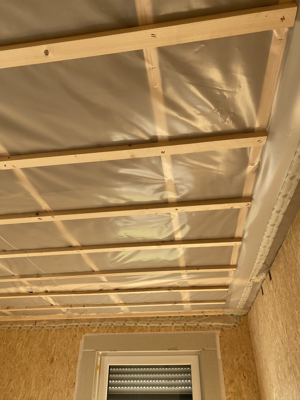 Open ceiling with wooden battens, foil insulation, and window at the lower section.
