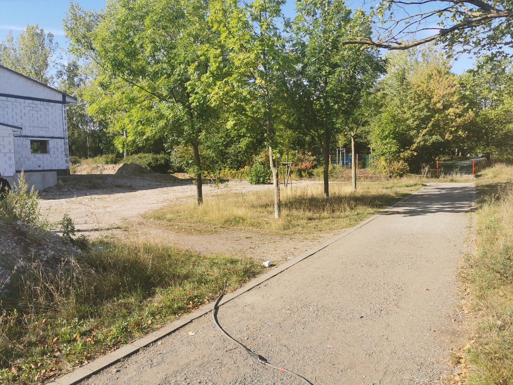 Unfertiger Rohbau links, gepflasterter Weg rechts, grüne Bäume, sonniger Tag.