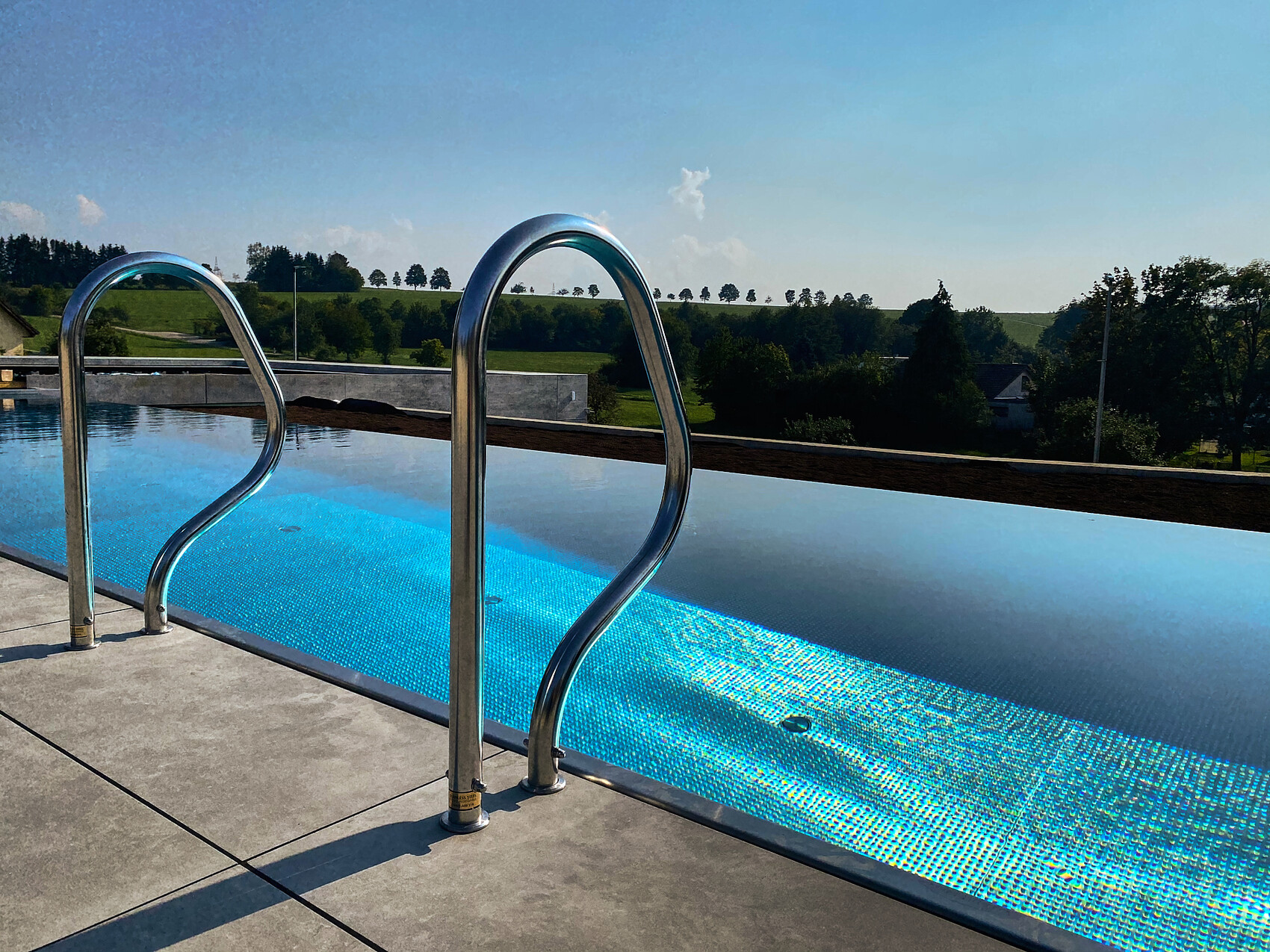 Outdoor pool with stainless steel handrails, turquoise water, green landscape in the background.