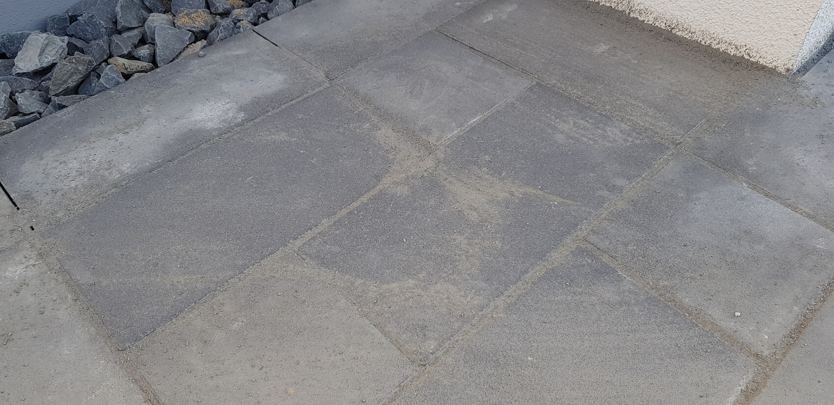 Paved concrete slabs on walkway, dust on the surface, stones in left corner, wall on right.