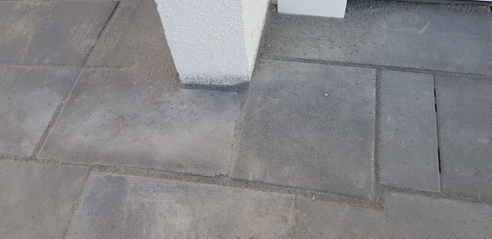 Paved walkway made of gray concrete slabs, near a white column base, dusty ground.