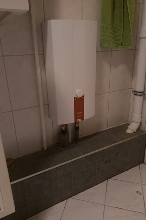 Water boiler on tiled wall with pipes underneath, next to a green towel rail.