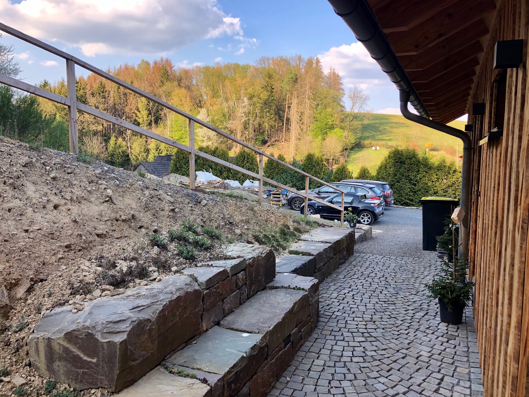 Footpath next to wooden house with stone steps, railing, cars, and forest in the background.