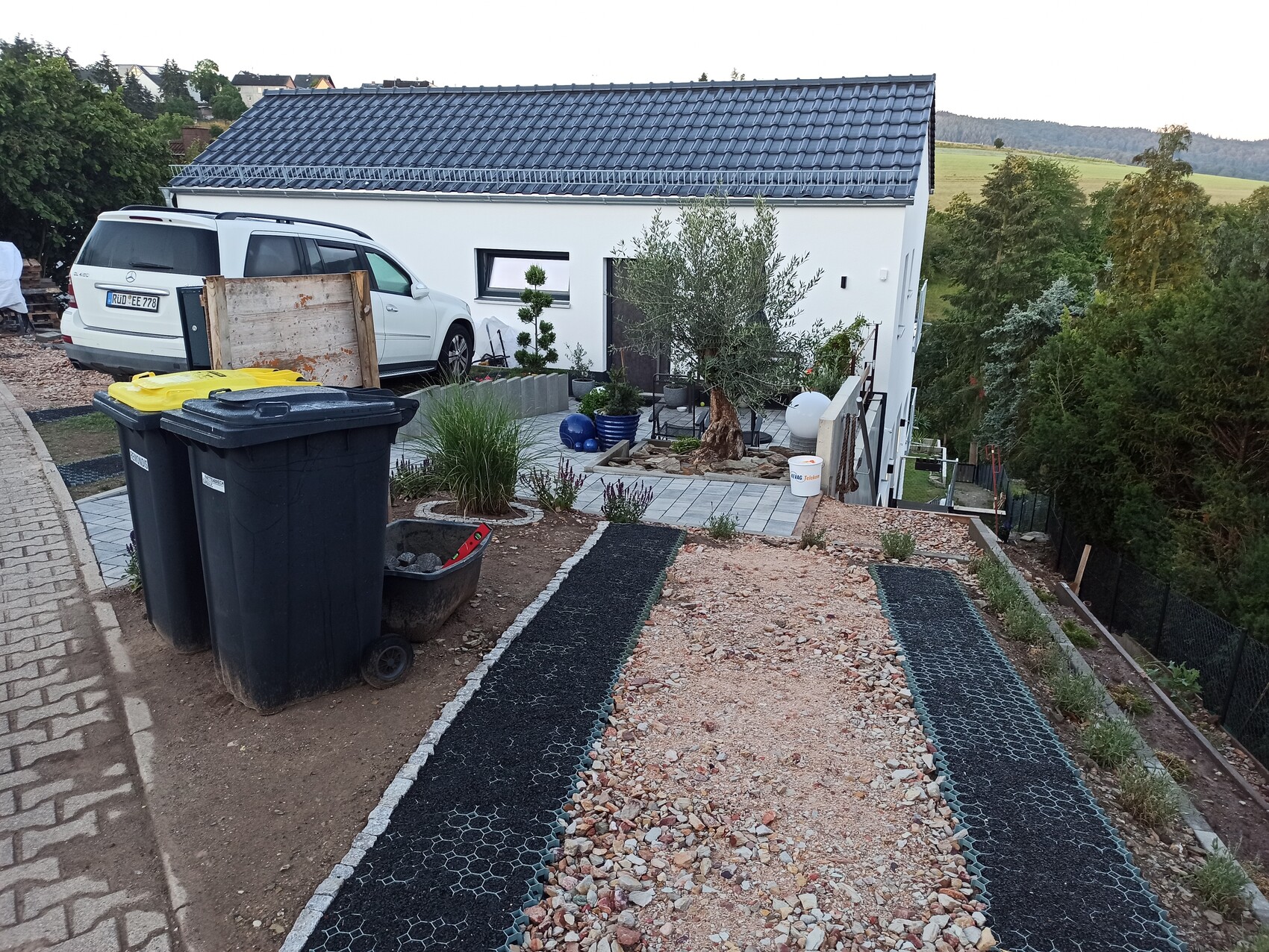 Modern white house with grey roof; courtyard with gravel path, trash bins, and car.